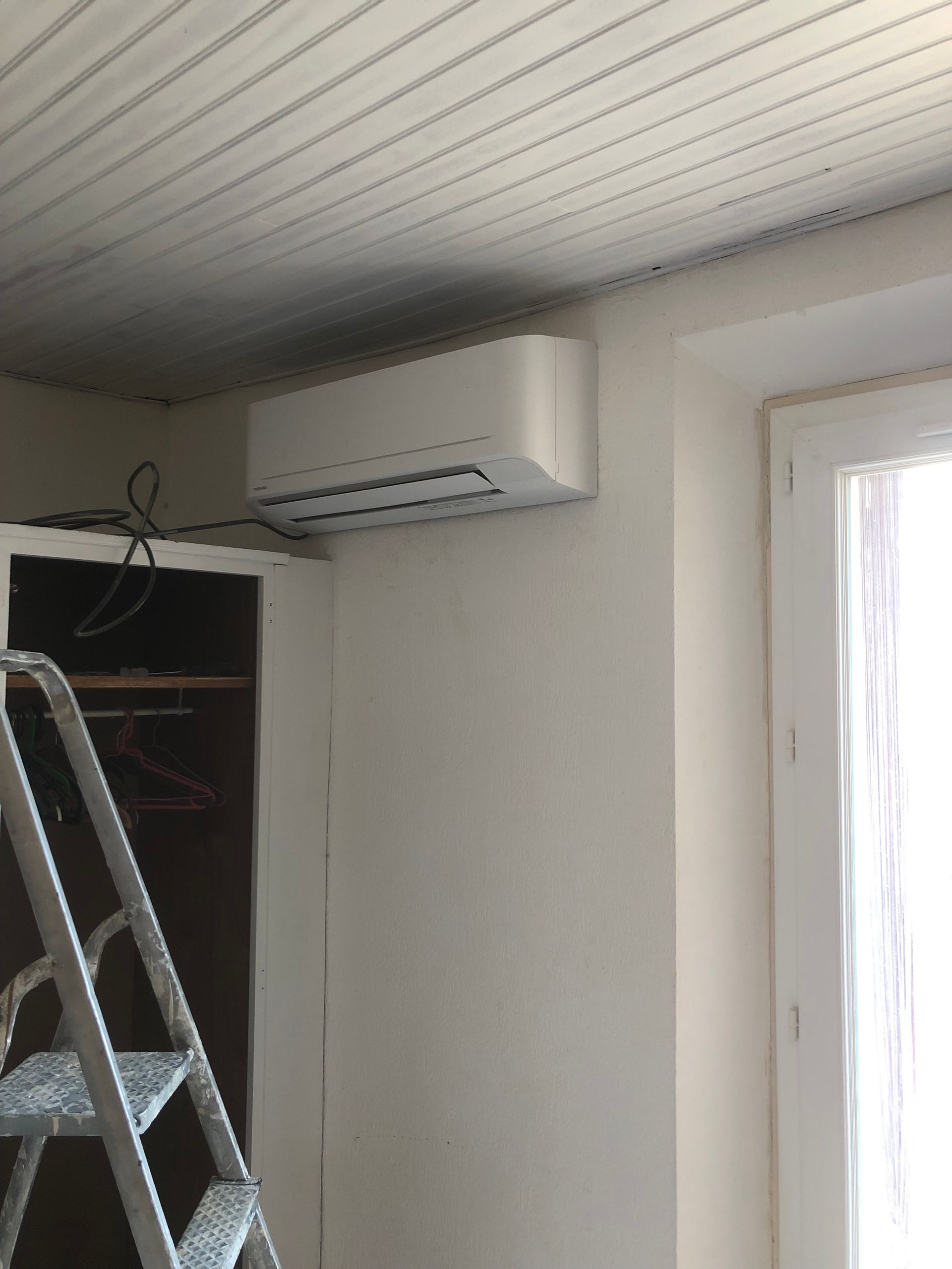Climatiseur blanc fixé sur un mur blanc, à côté d'un placard et d'une fenêtre. Une échelle est visible au premier plan.