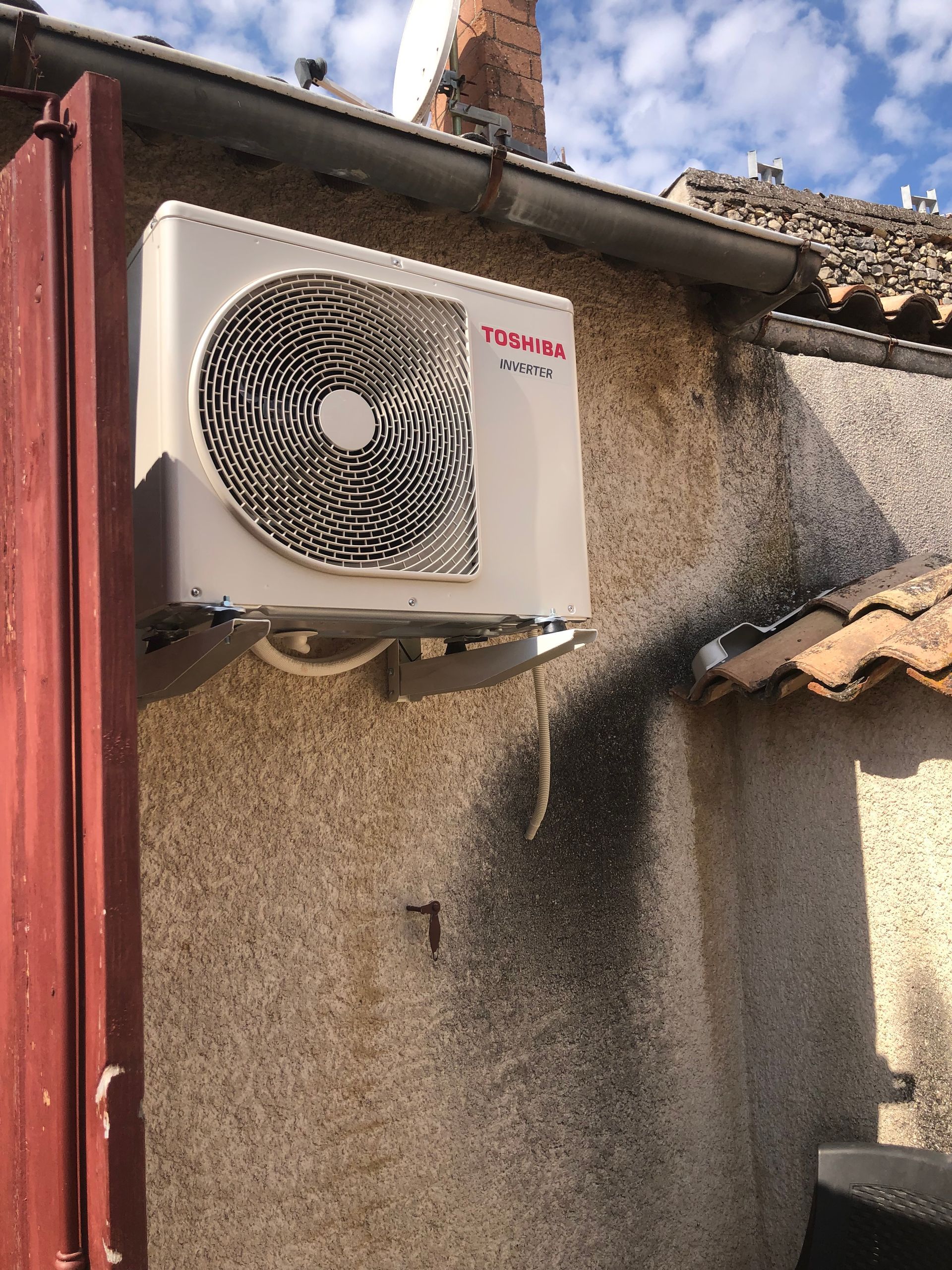 Une unité de climatisation extérieure montée sur un mur en stuc patiné, près d'un toit en tuiles.