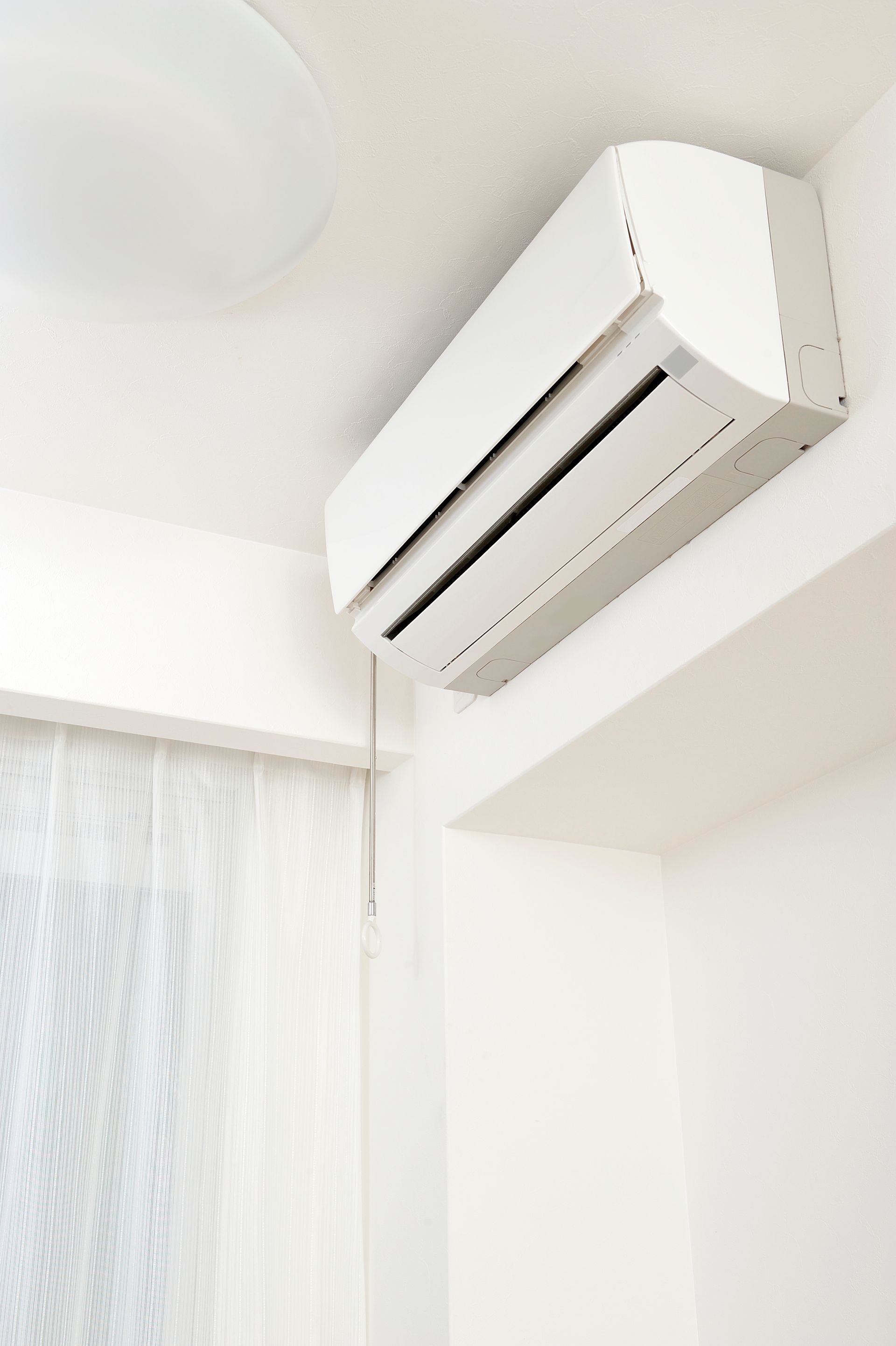 Climatiseur blanc monté sur un mur blanc à côté d'une fenêtre avec un rideau transparent.