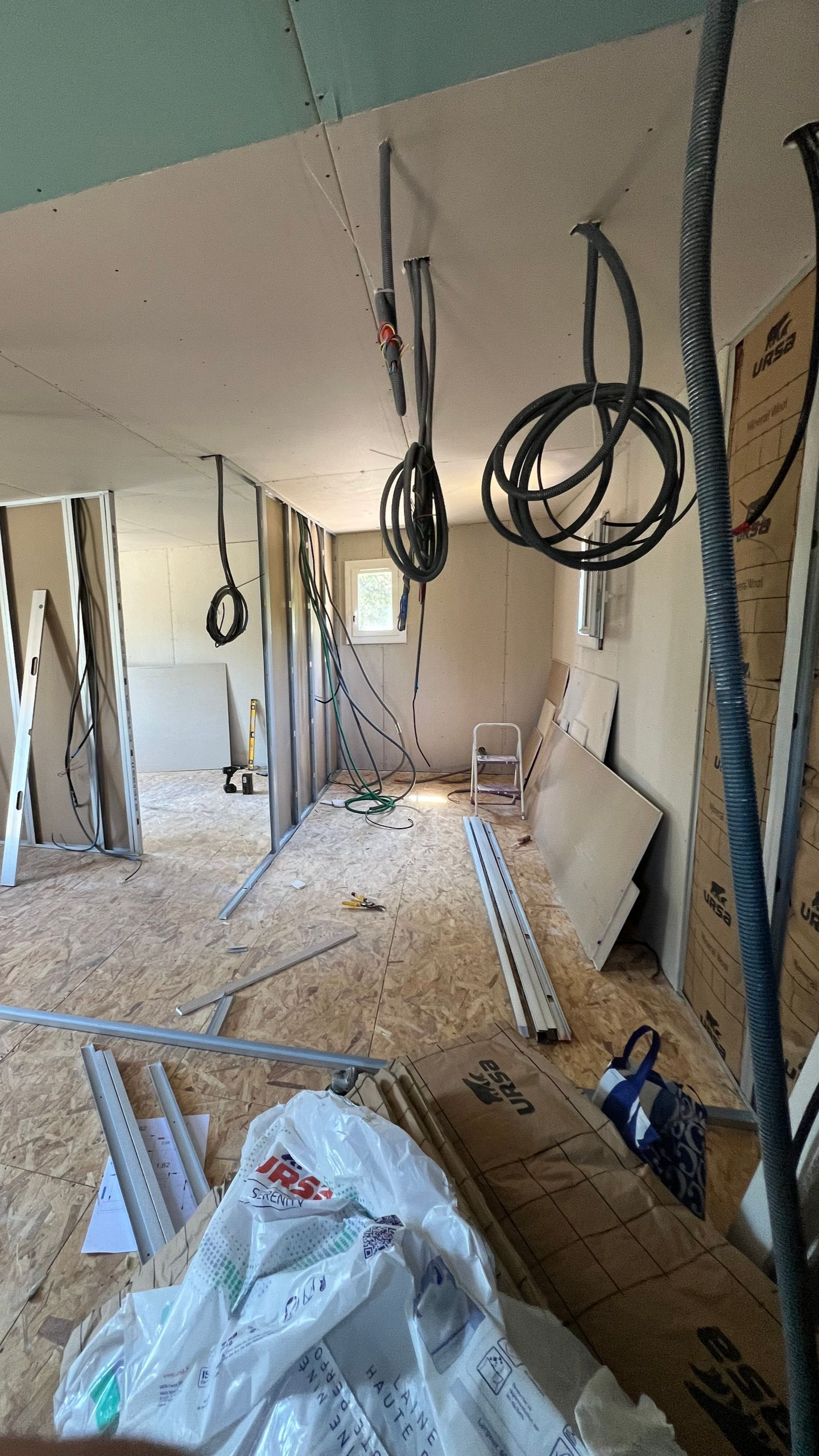 Intérieur d'une pièce en construction avec câblage exposé, murs en contreplaqué et plafond partiel en cloison sèche.