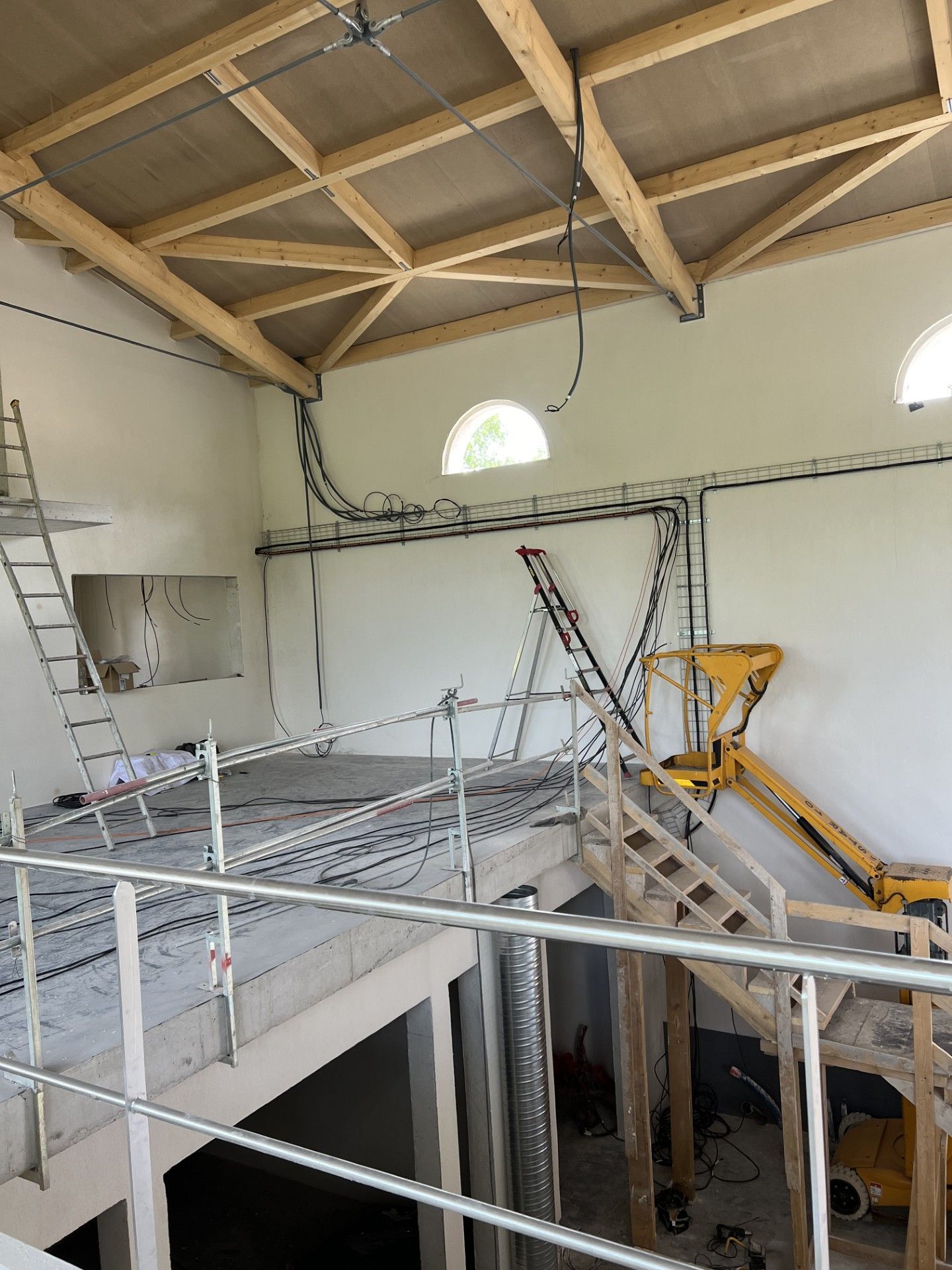 Vue intérieure d'un bâtiment en construction avec poutres en bois apparentes, conduits et mezzanine partielle.