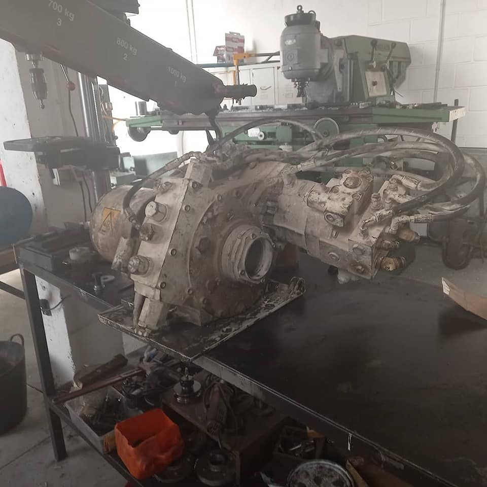 Un componente mecánico grande y sucio sobre una mesa de trabajo, con equipos de taller al fondo.