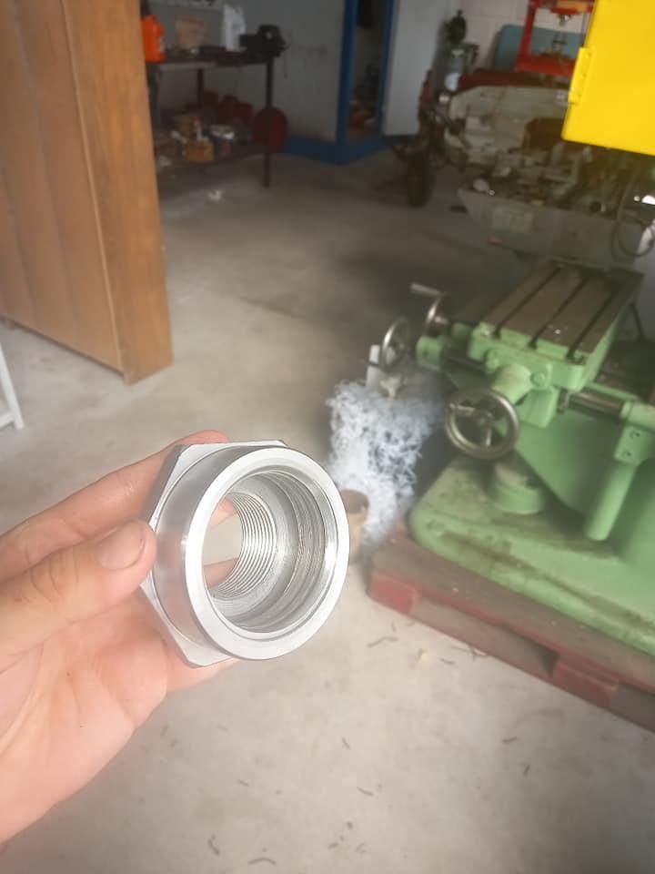 Una mano sostiene un herraje metálico roscado de plata. Al fondo, se ven herramientas y equipos de taller.