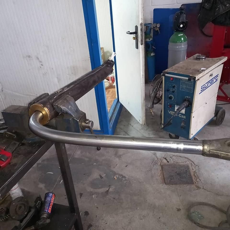 En un taller se dobla un tubo de metal; al fondo hay una máquina de soldar y otras herramientas.