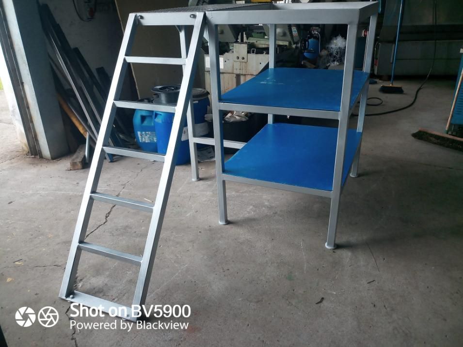 Mesa de trabajo de metal con estantes azules y escalera adjunta, en el interior sobre un piso de concreto.