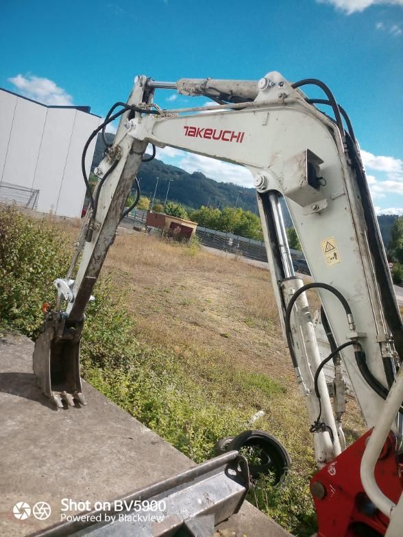 Excavadora Takeuchi blanca y gris sobre una plataforma de hormigón con una zona de césped al fondo y cielo azul.