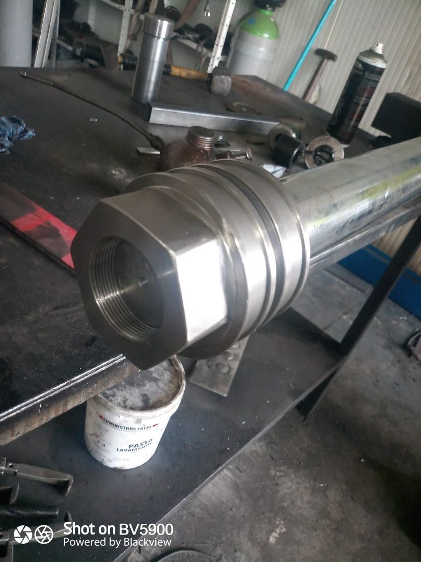 Cilindro metálico con extremo hexagonal, probablemente parte de un sistema hidráulico, apoyado sobre una mesa de taller.