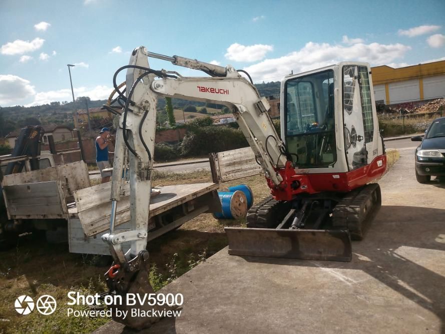 Una pequeña excavadora blanca y roja estacionada al lado de un camión de plataforma sobre concreto.