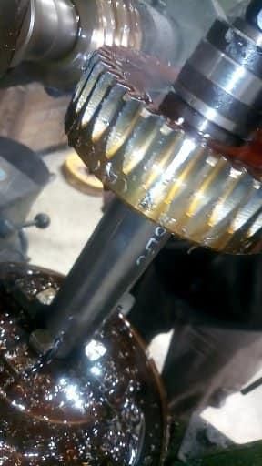 Una máquina cortando dientes en un engranaje grande de metal, cubierto de aceite, con una broca visible.