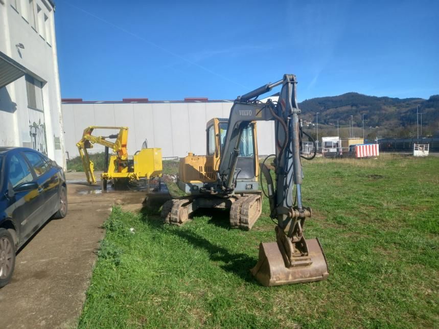 Excavadora amarilla y maquinaria sobre el césped cerca de un edificio en un día soleado.