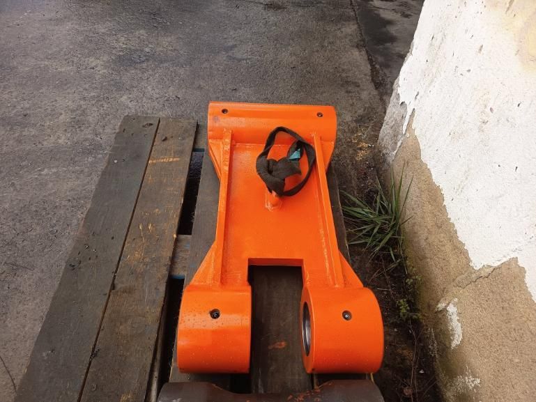 Una pieza metálica de color naranja con extremos cilíndricos reposa sobre una paleta de madera junto a una pared blanca.