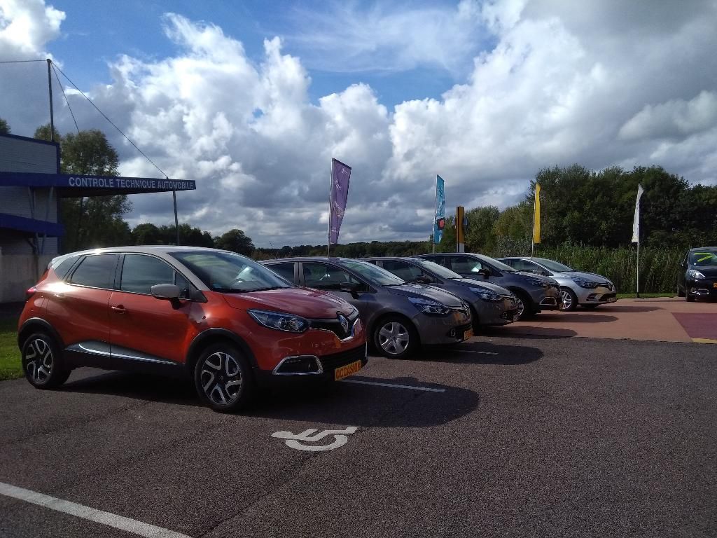 Une rangée de voitures garées, menée par un SUV orange, sur un parking ensoleillé.