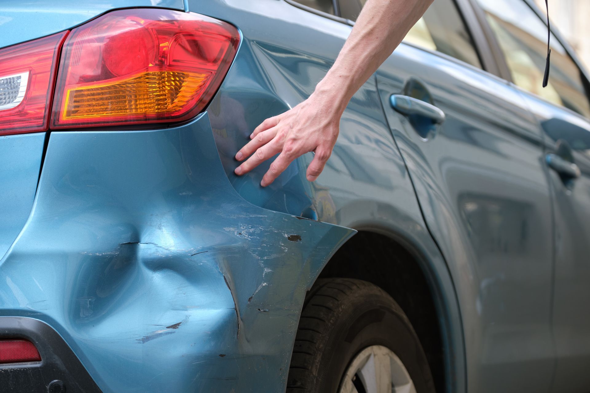 Main touchant le panneau latéral arrière cabossé d'une voiture bleue, près du feu arrière.