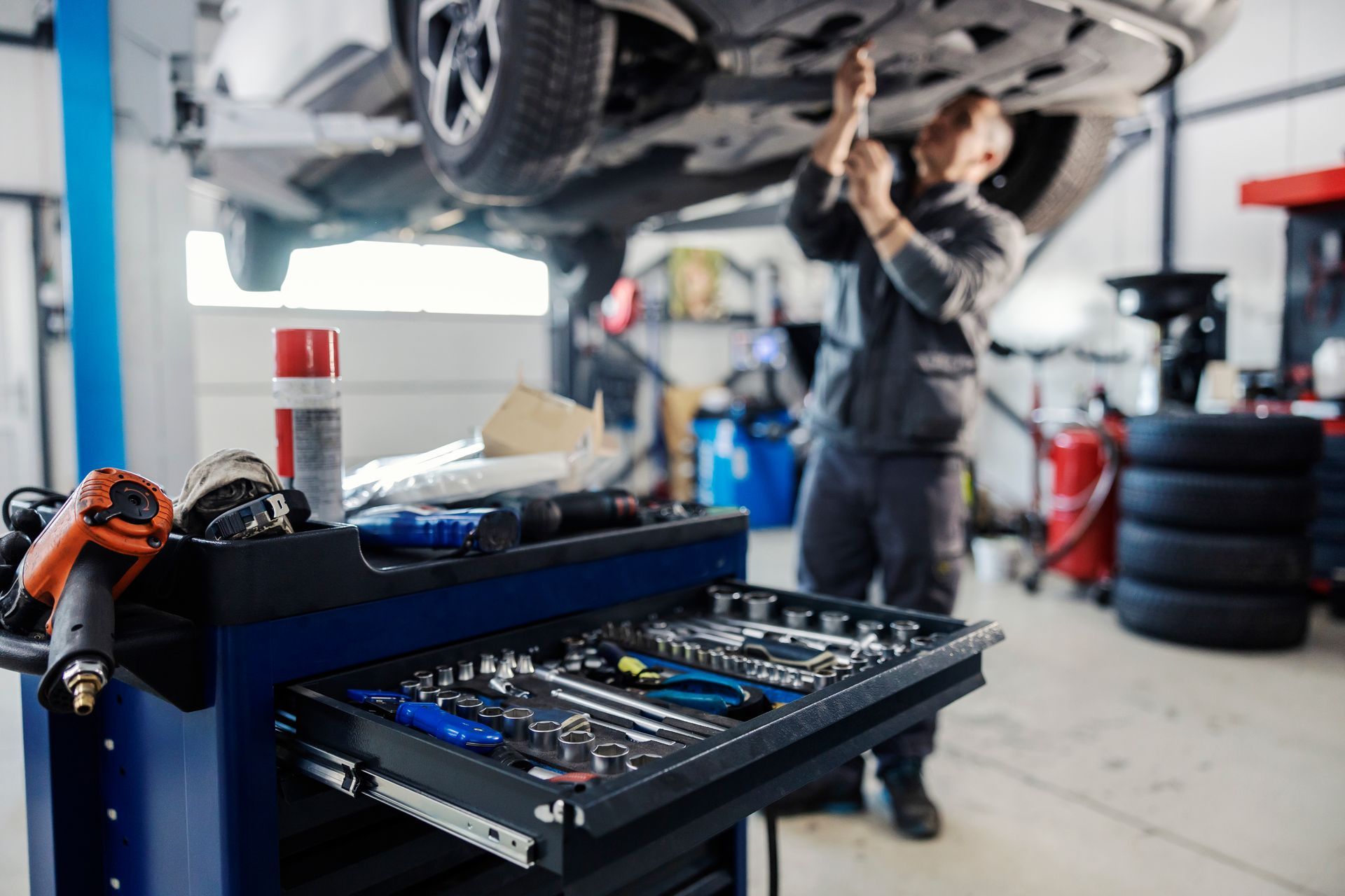 Un mécanicien travaille sous une voiture dans un garage avec un chariot à outils ouvert au premier plan.