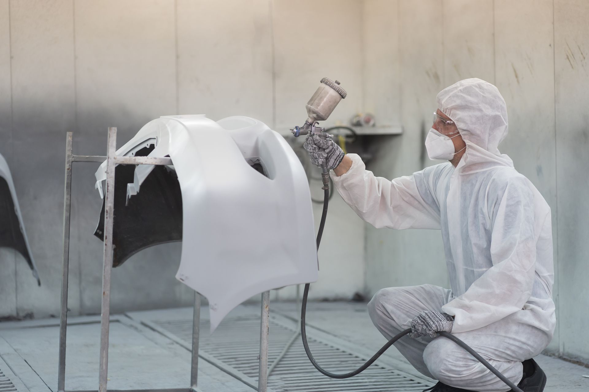 Une personne en combinaison de protection blanche peint à la bombe la carrosserie d'une voiture dans une cabine de peinture.