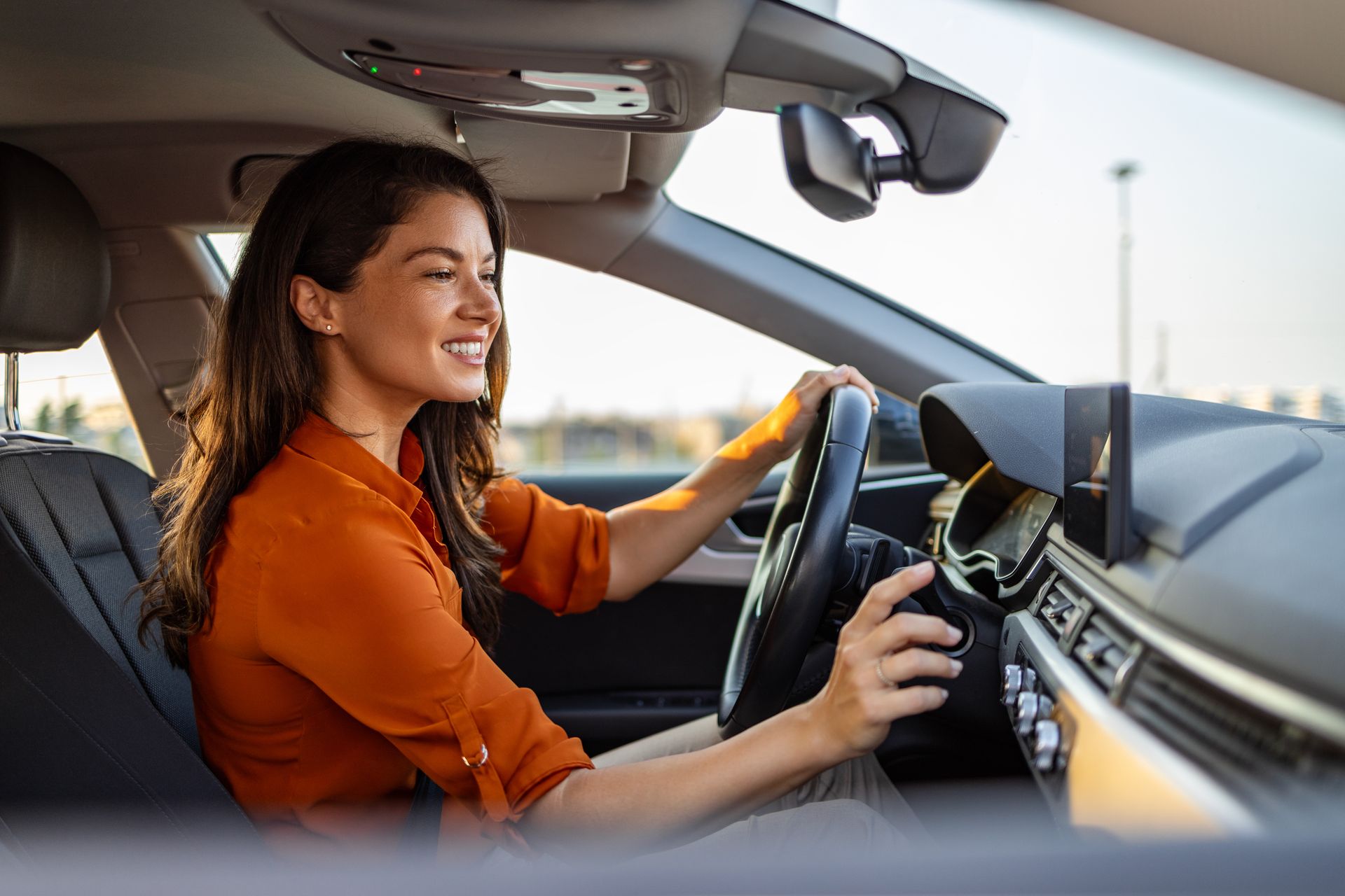 Une femme conduit une voiture, souriante, une main sur le volant et le tableau de bord visible.