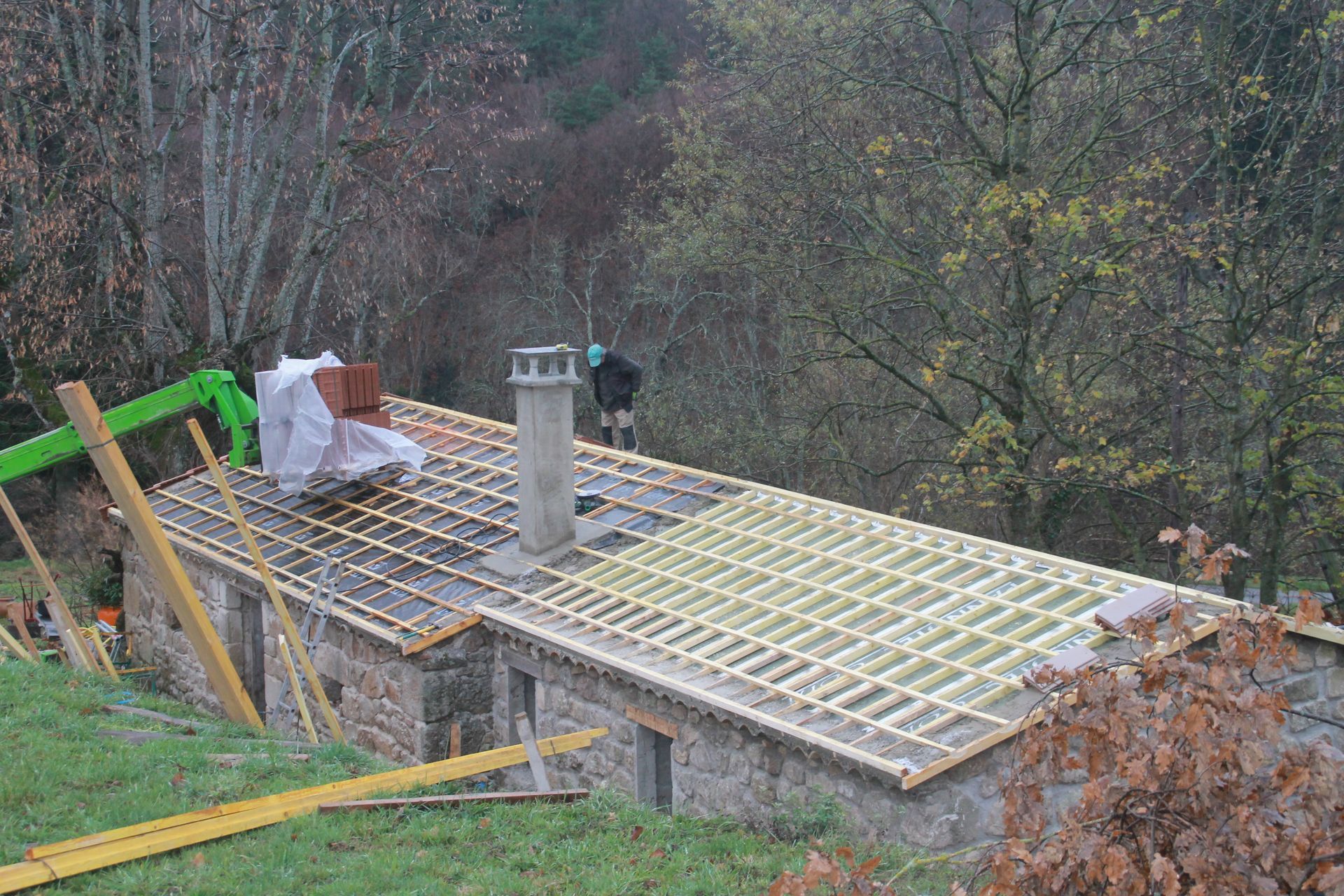 Toiture en construction à flanc de colline boisée, avec une charpente apparente et une cheminée.
