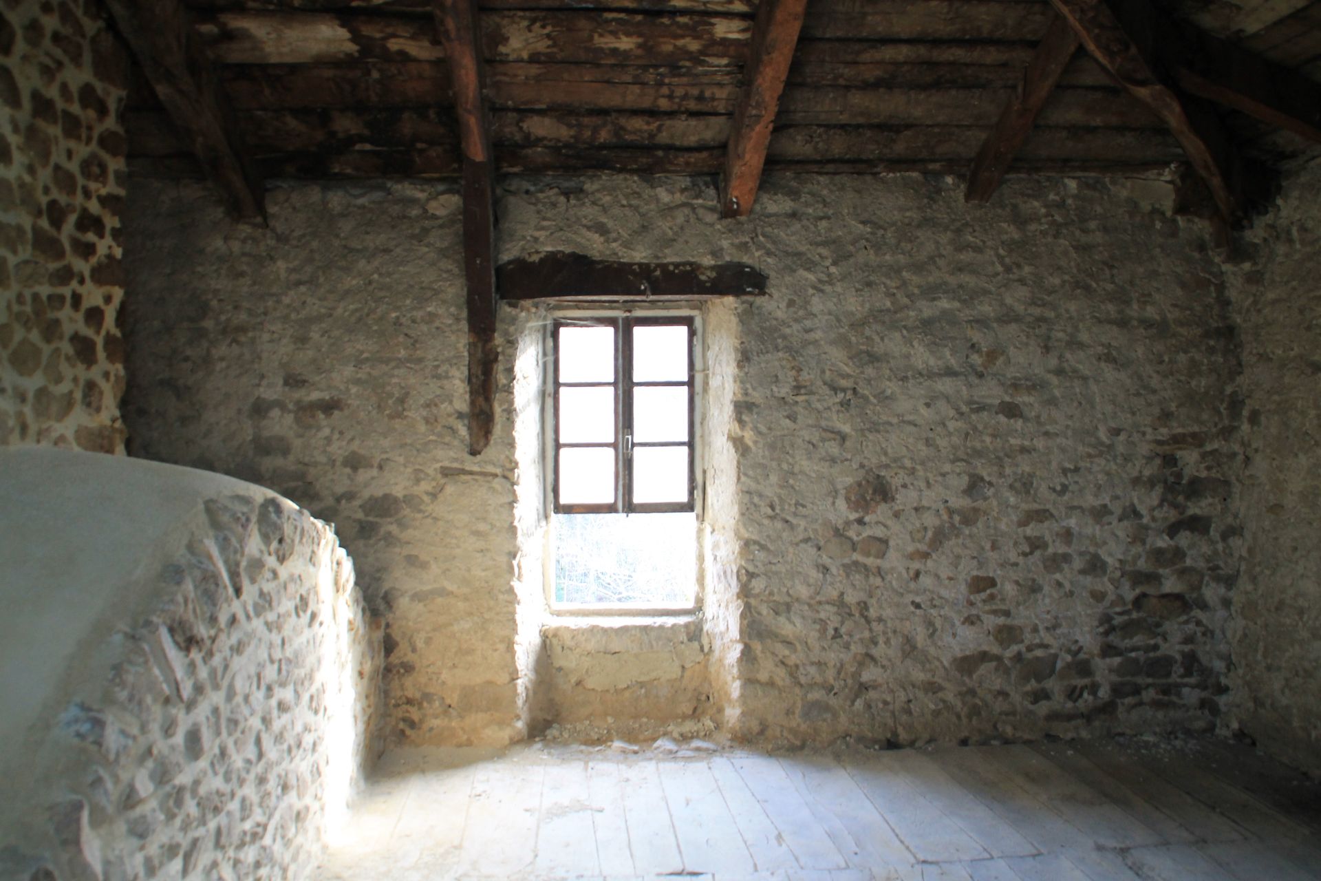 Pièce mansardée en pierre avec poutres apparentes et une petite fenêtre laissant entrer la lumière