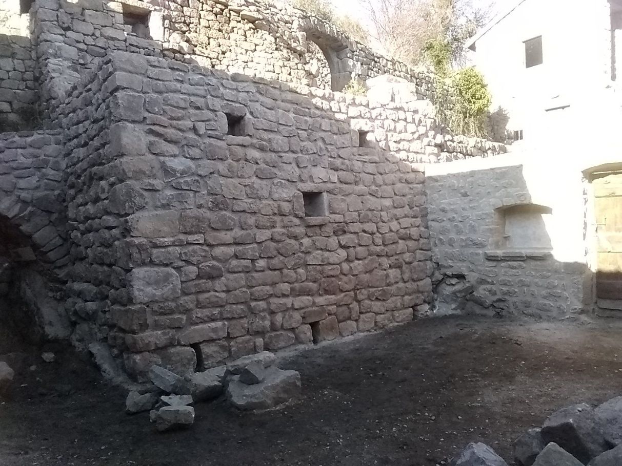 Cour en pierre avec un mur de soutènement brut et des façades baignées de soleil.