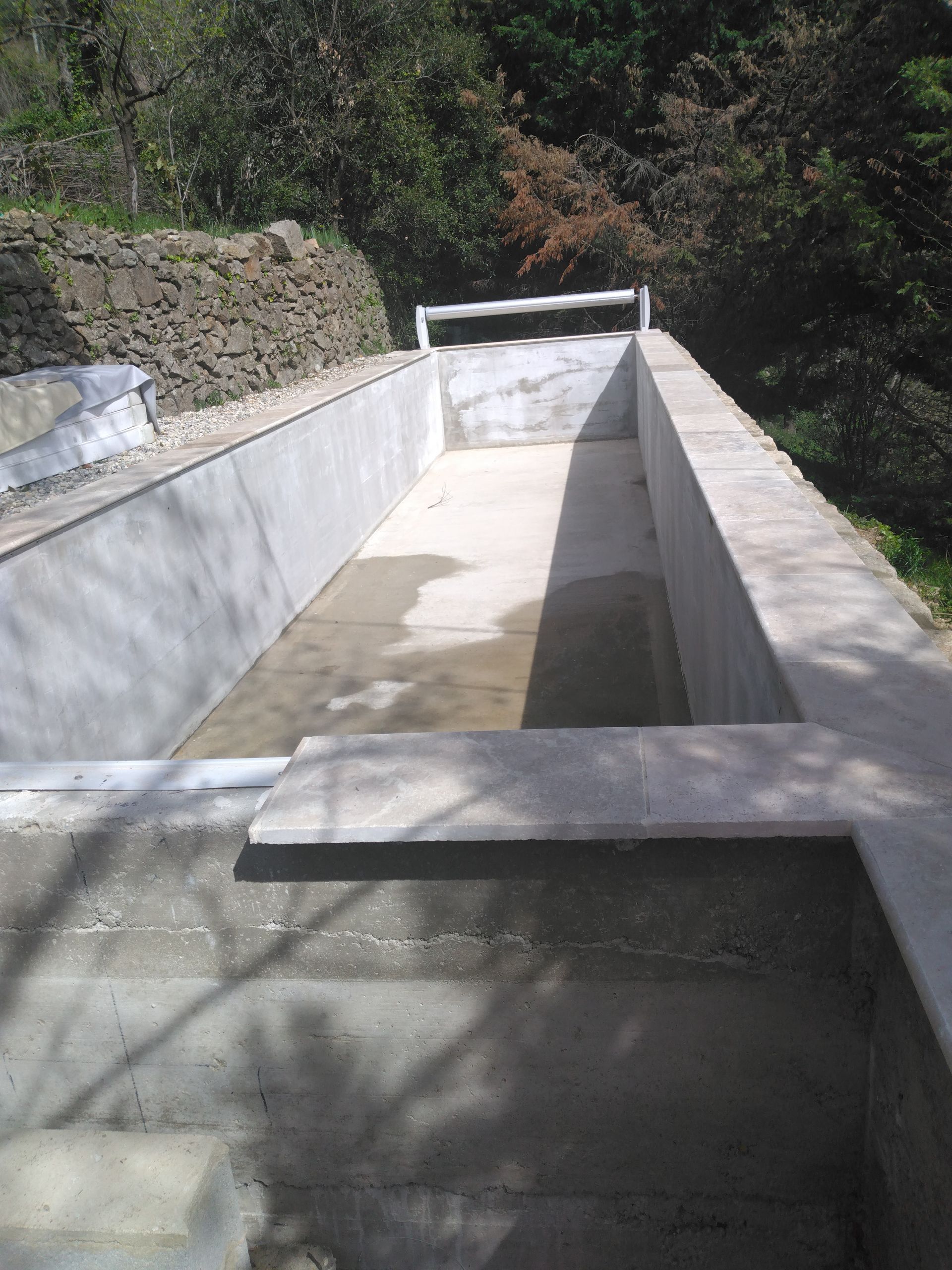 Étroit canal de drainage en béton descendant la pente le long d'un mur de soutènement en pierre.