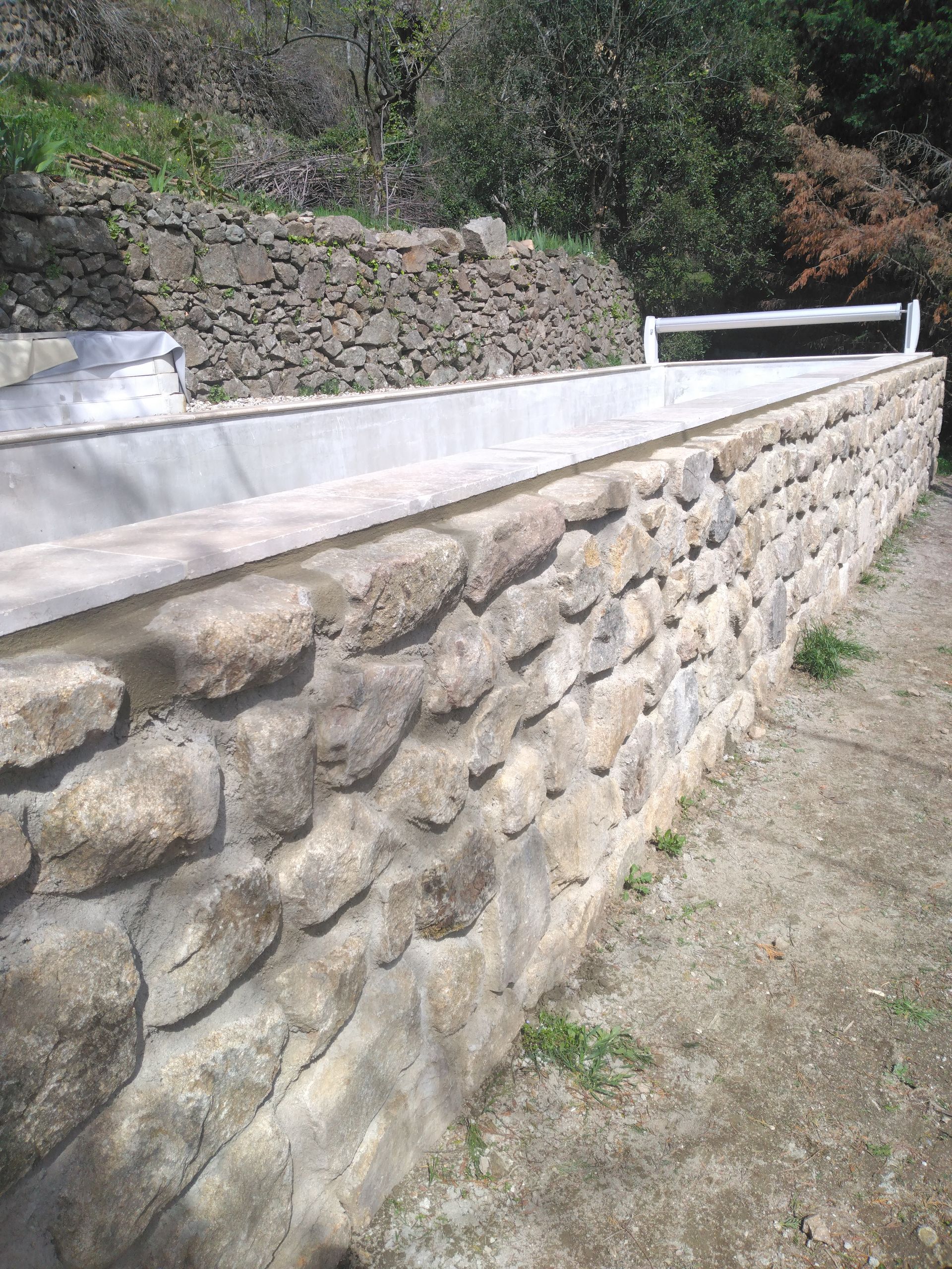 Mur de soutènement en pierre bordant un chemin de terre, avec des arbres verts en arrière-plan.
