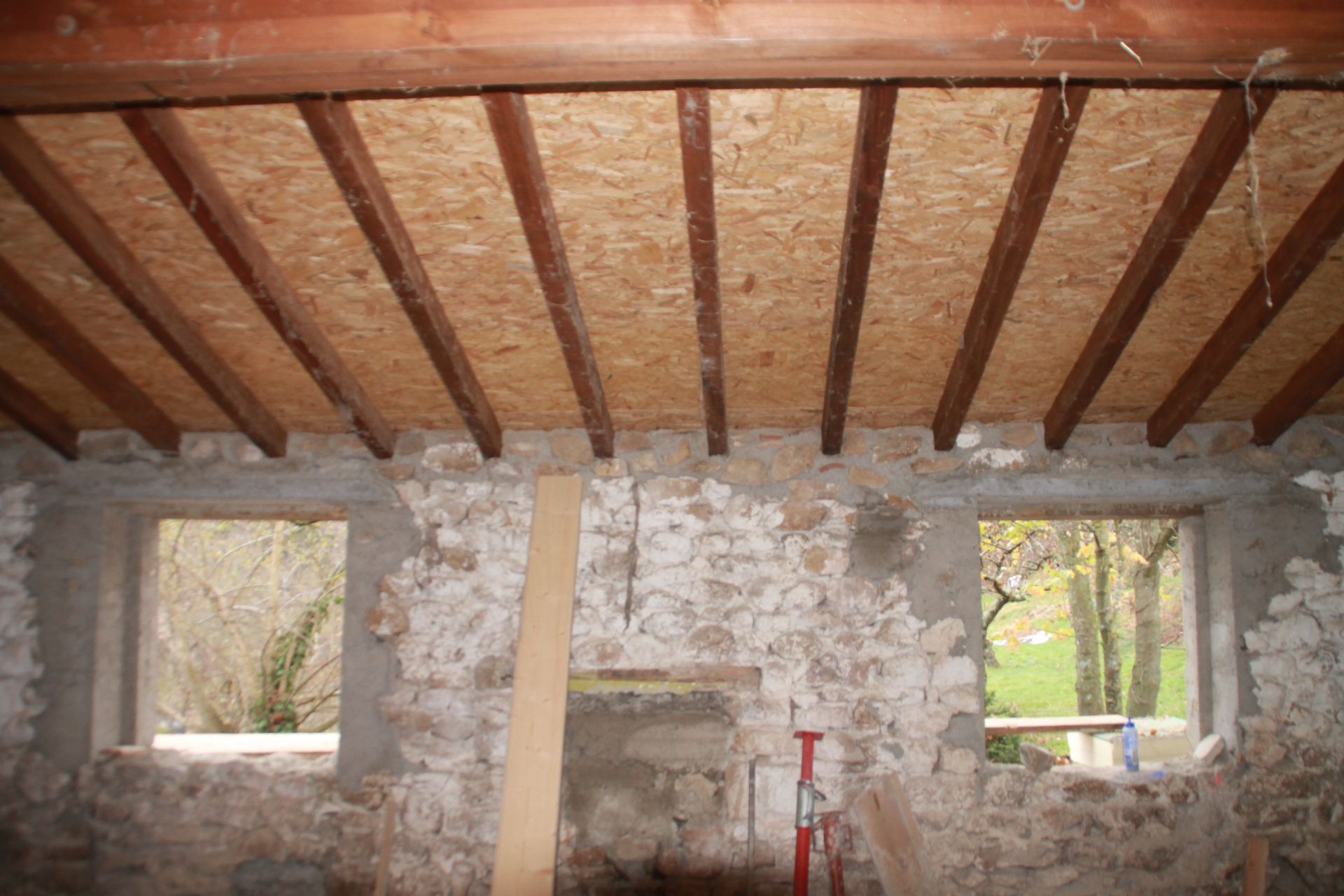 Poutres apparentes au plafond, surplombant des murs en pierre brute, dans une pièce rustique avec des ouvertures de fenêtres.