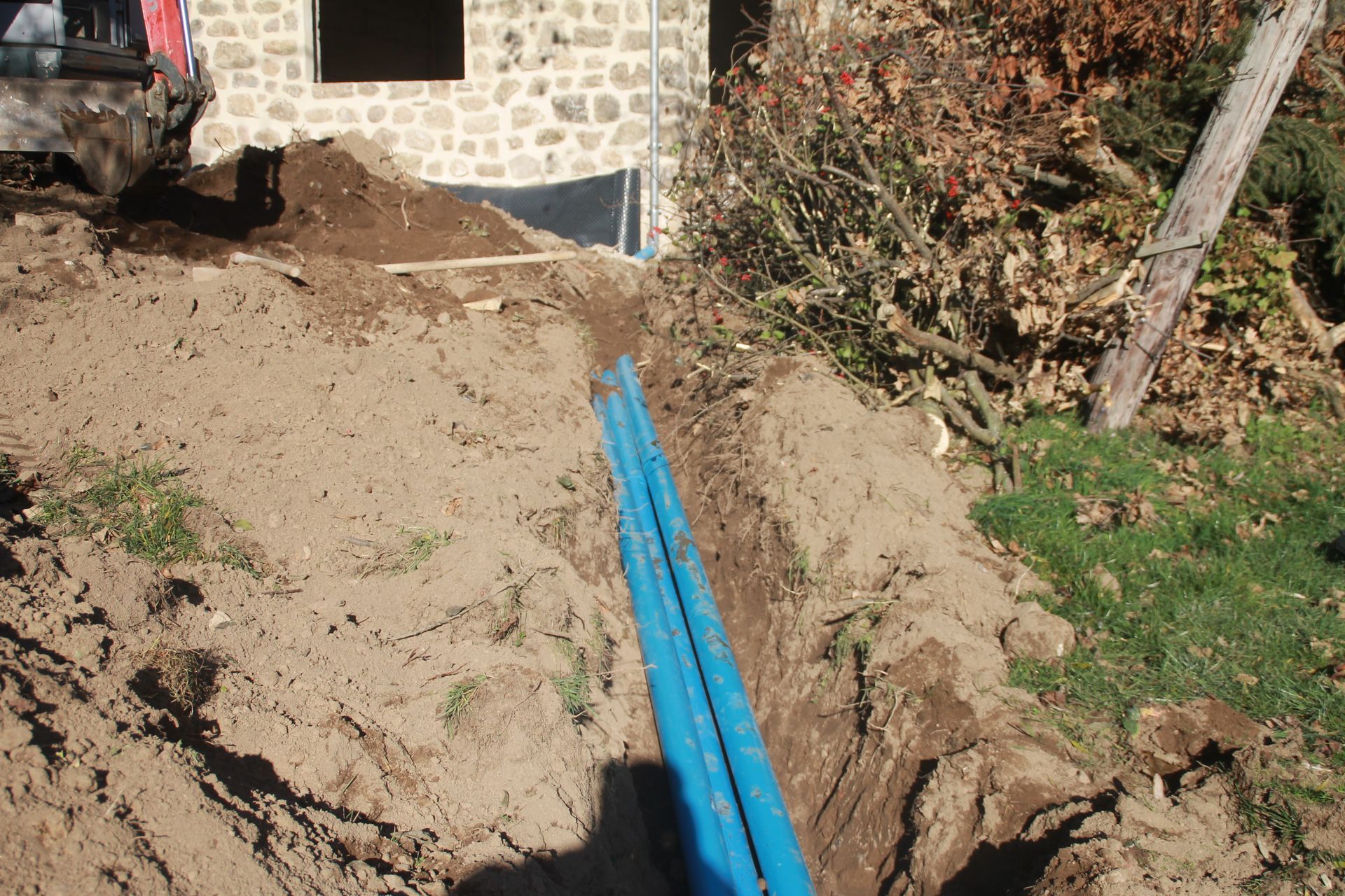 Des tuyaux bleus posés dans une tranchée de terre à côté d'une maison en pierre et d'un chemin de jardin.