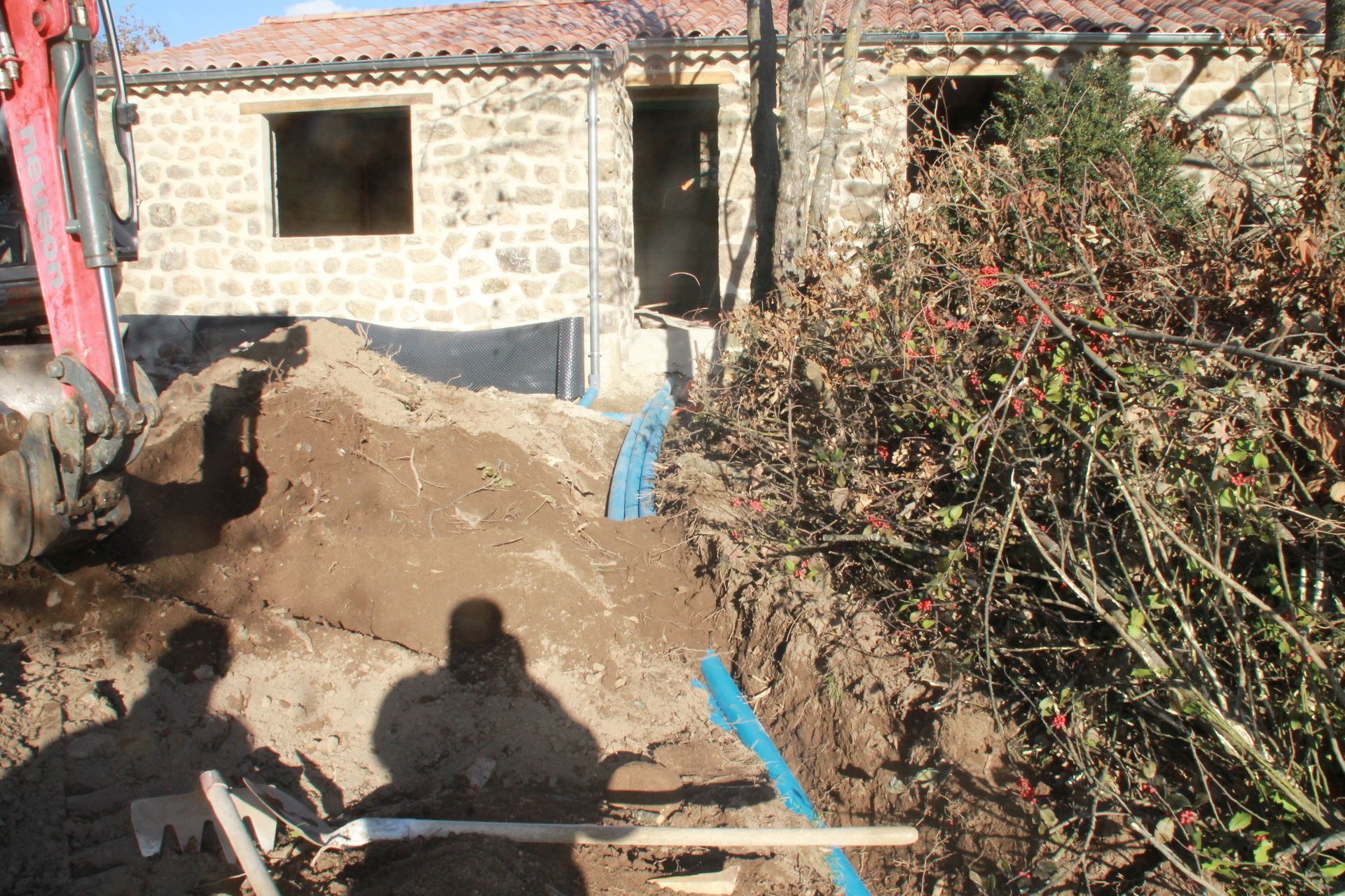 Maison en pierre en construction, avec des tas de terre, des tuyaux bleus et un buisson feuillu devant.