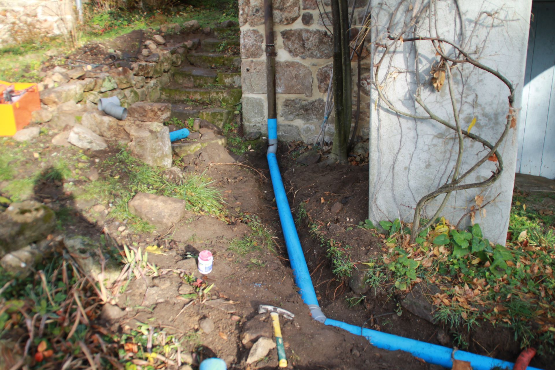 Un tuyau bleu longe un chemin de terre, à côté d'un mur en béton et de marches en pierre, dans une cour extérieure.