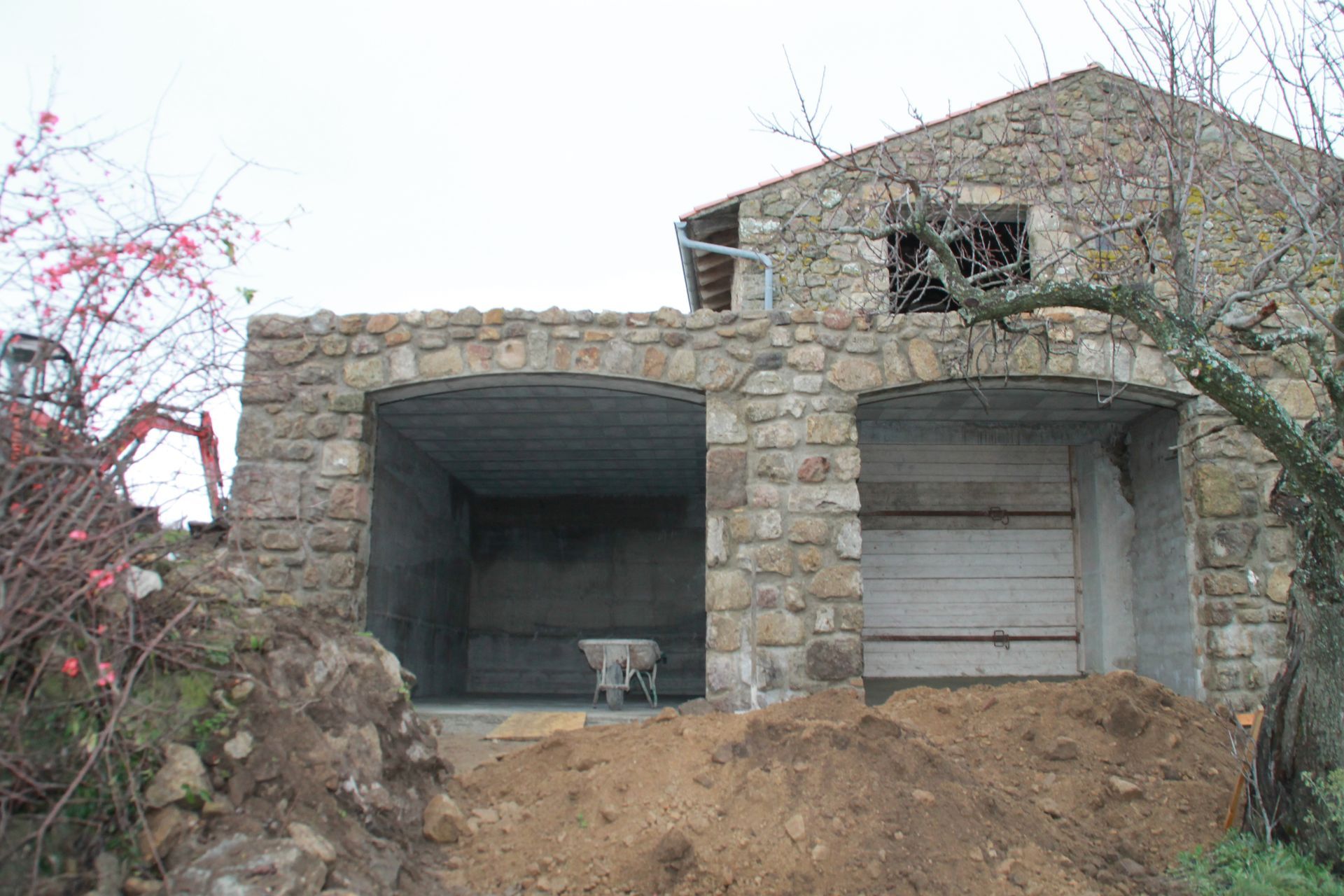 Maison en pierre en construction avec deux garages ouverts et un terrain en terre devant.
