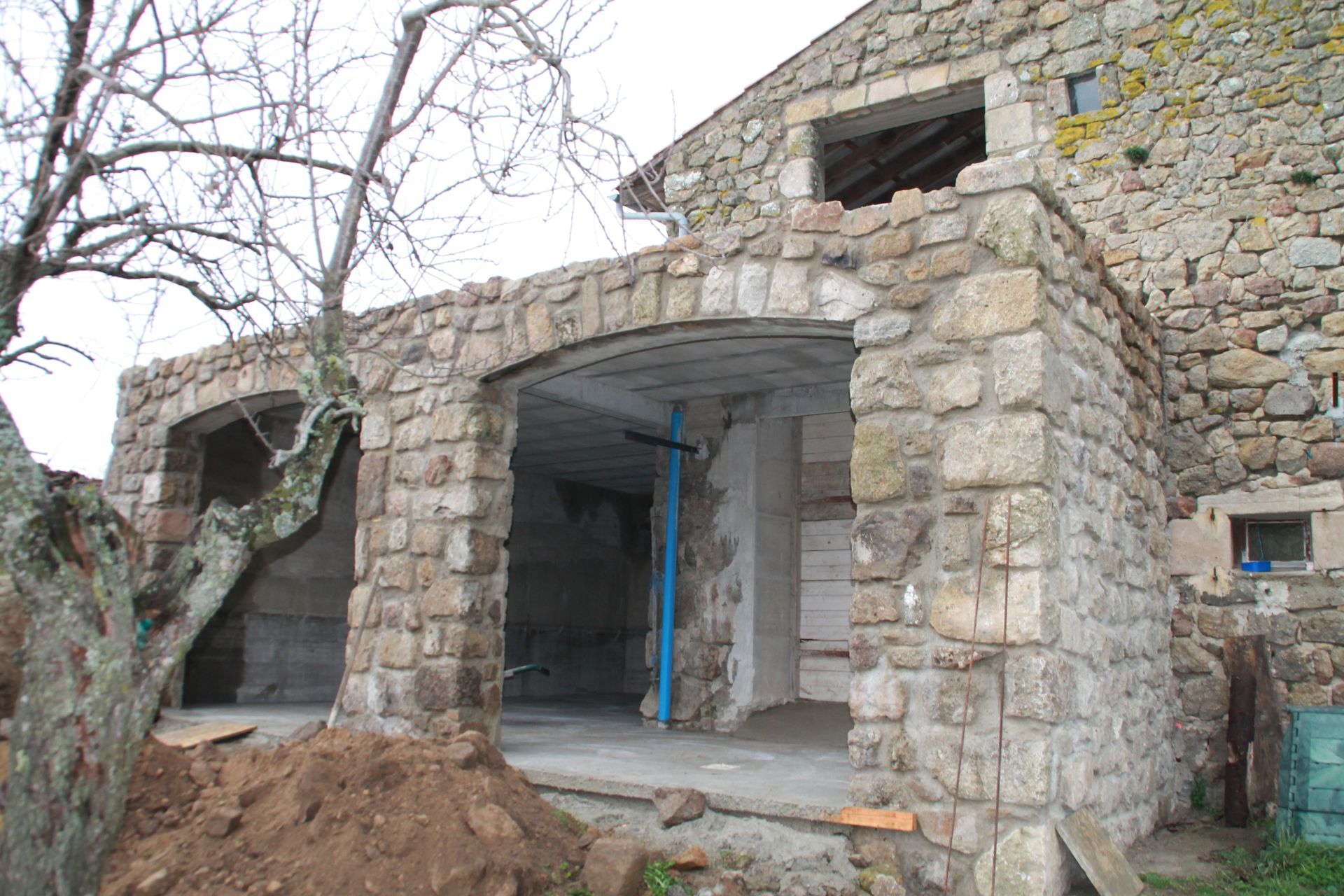 Bâtiment en pierre en construction, avec une porte ouverte et un arbre nu devant.