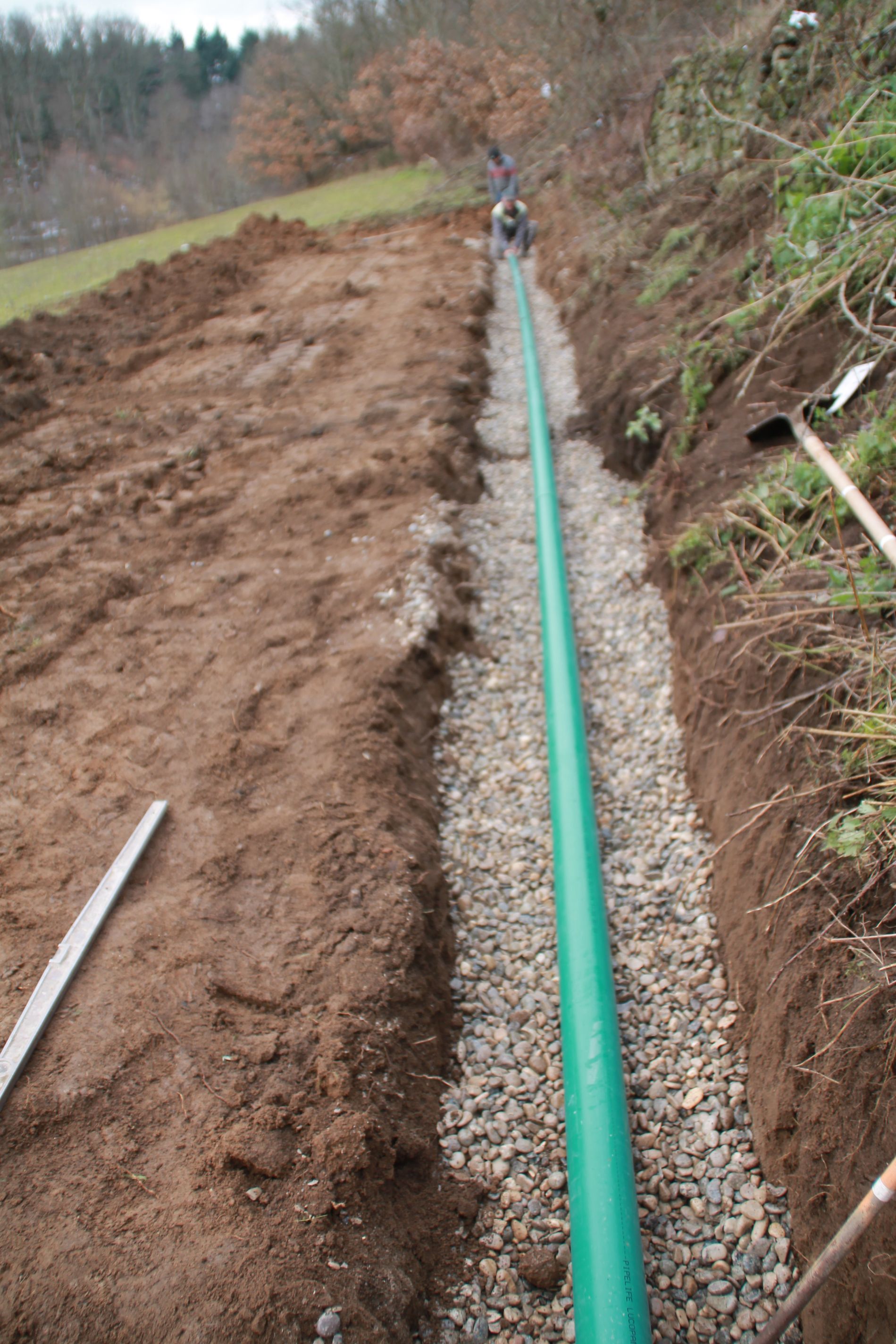 Un tuyau vert est installé dans une tranchée au bord d'un chemin de terre boueux et d'une pente herbeuse.