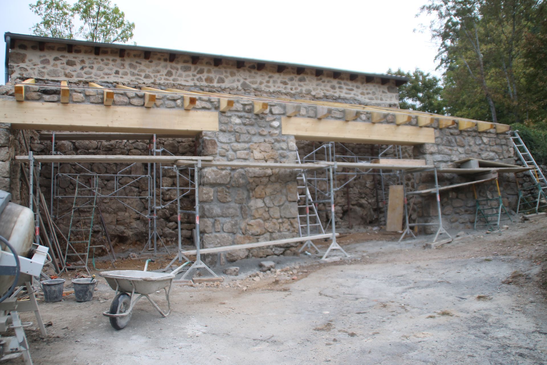 Bâtiment en pierre en construction avec des échafaudages, une brouette et des ouvertures murales inachevées.