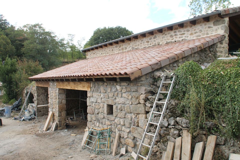 Maison en pierre en construction avec un toit de tuiles rouges, une échelle en bois et de la végétation environnante.