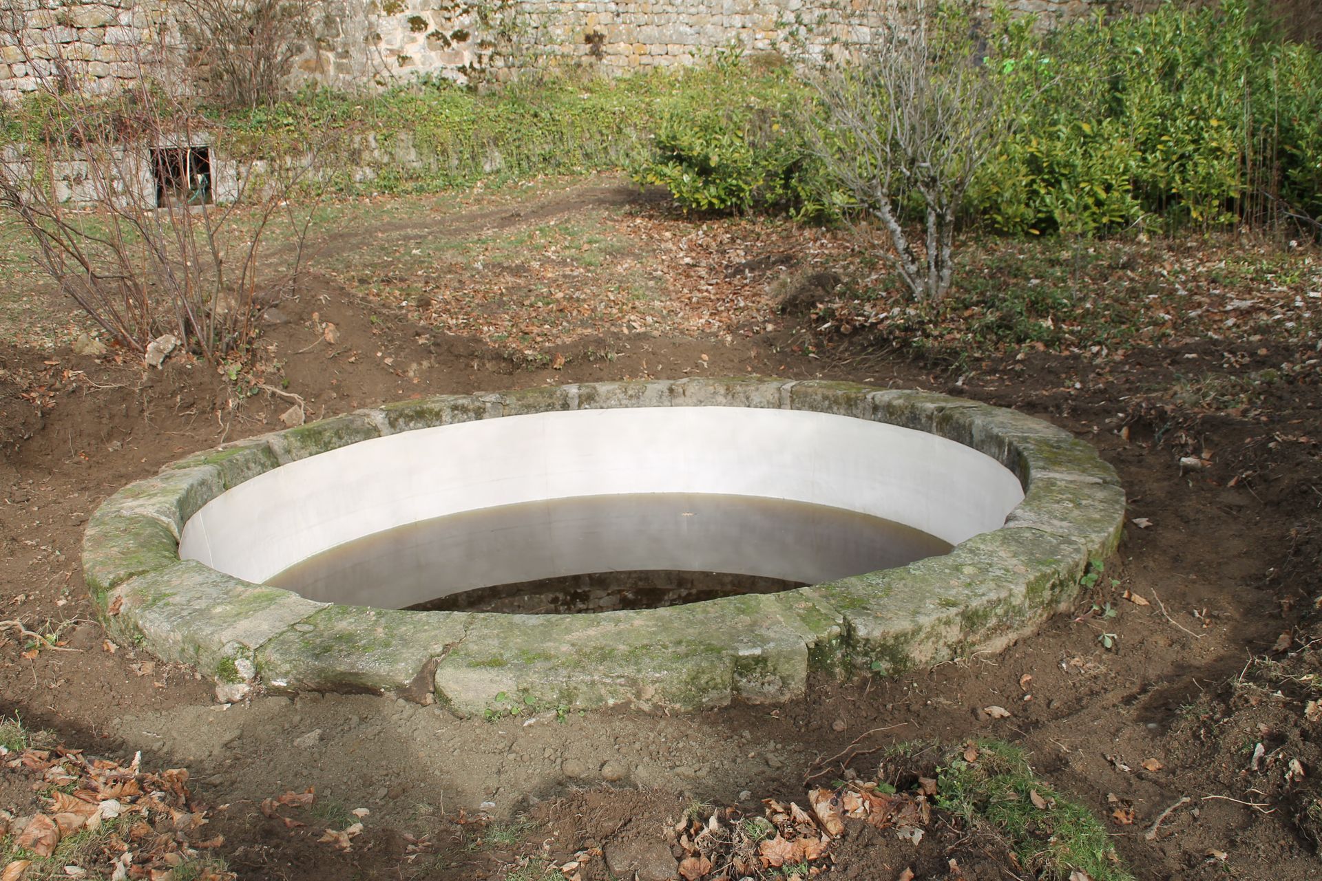 Un vieux puits en pierre dans une clairière de terre, près d'un mur moussu et d'arbustes.