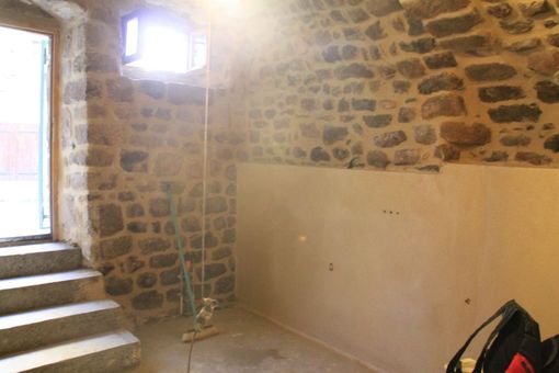 Pièce aux murs de pierre avec une fenêtre lumineuse, des marches sur la gauche et un panneau mural peint en beige.