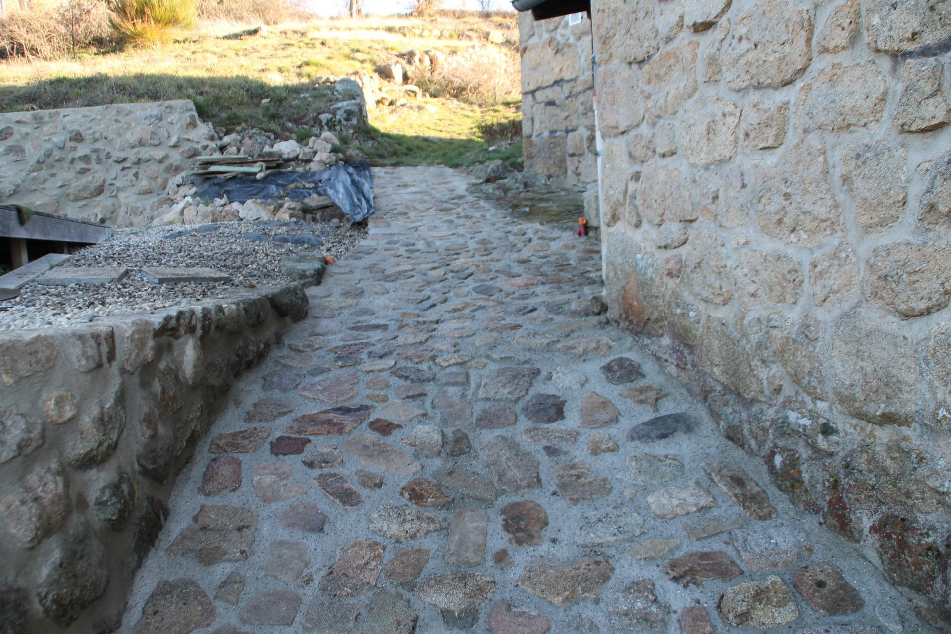 Chemin pavé longeant un mur de pierre, menant à un espace extérieur ensoleillé.