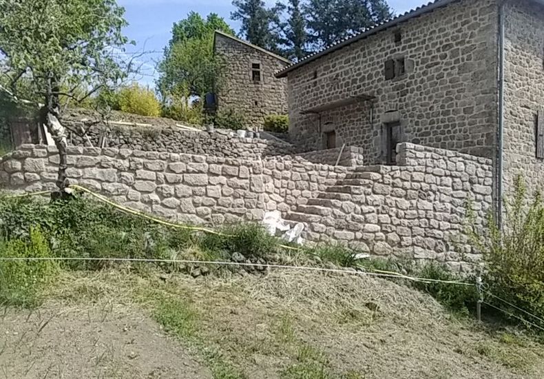 Maison en pierre avec un mur de soutènement en pente sur un coteau herbeux sous les arbres et le ciel bleu