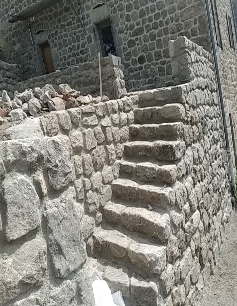 Escalier en pierre sur la façade rustique d'un bâtiment en pierre, avec une rampe en métal et des murs en maçonnerie brute.