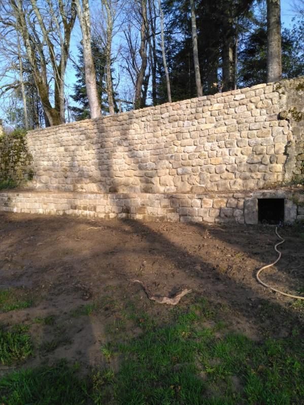 Mur de soutènement en pierre dans un jardin boisé, avec une petite ouverture en bas à droite.