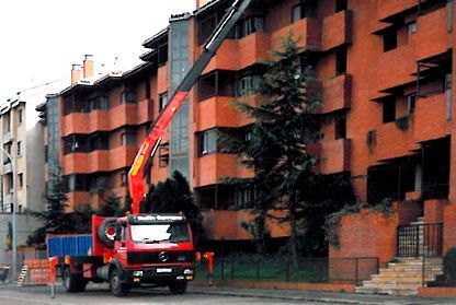 Camión de bomberos rojo con escalera elevada estacionado junto a un edificio de apartamentos de ladrillo naranja en una calle mojada.