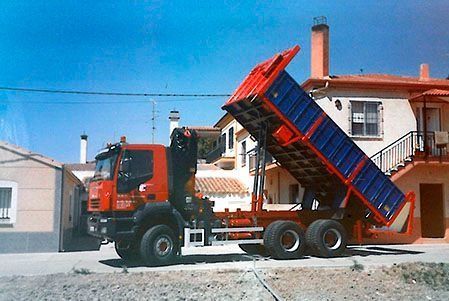 Un camión volquete naranja descarga un remolque azul con grava en el patio de una casa bajo un cielo despejado.