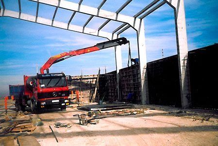 Un camión grúa de construcción rojo levanta vigas de acero dentro de la estructura de un edificio bajo un cielo azul.