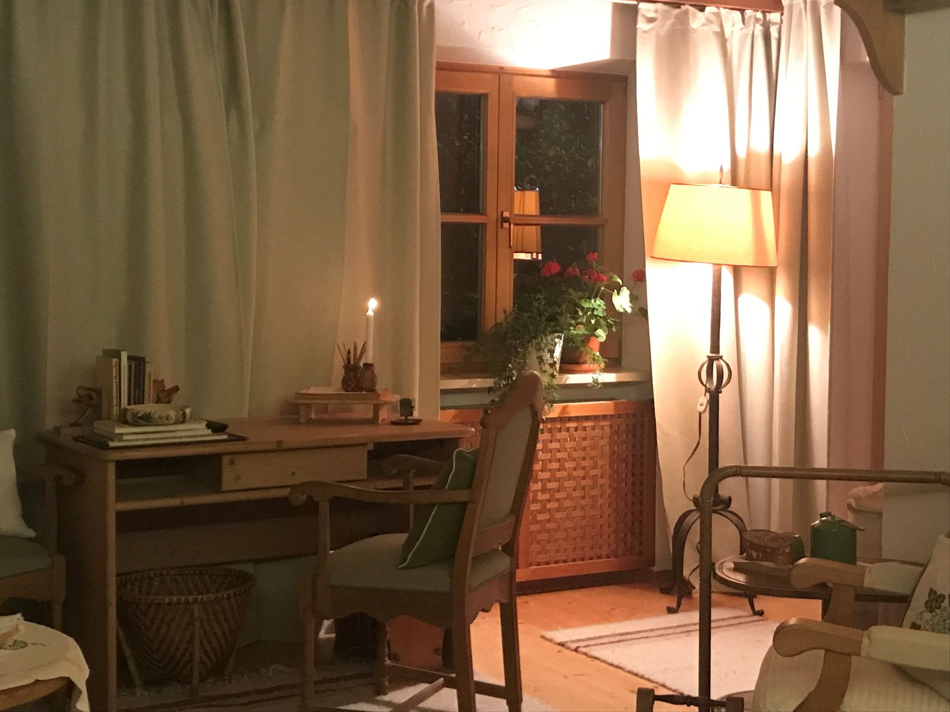 Gemütliches Zimmer mit Schreibtisch am Fenster, beleuchtet von einer Lampe. Hellfarbene Vorhänge.