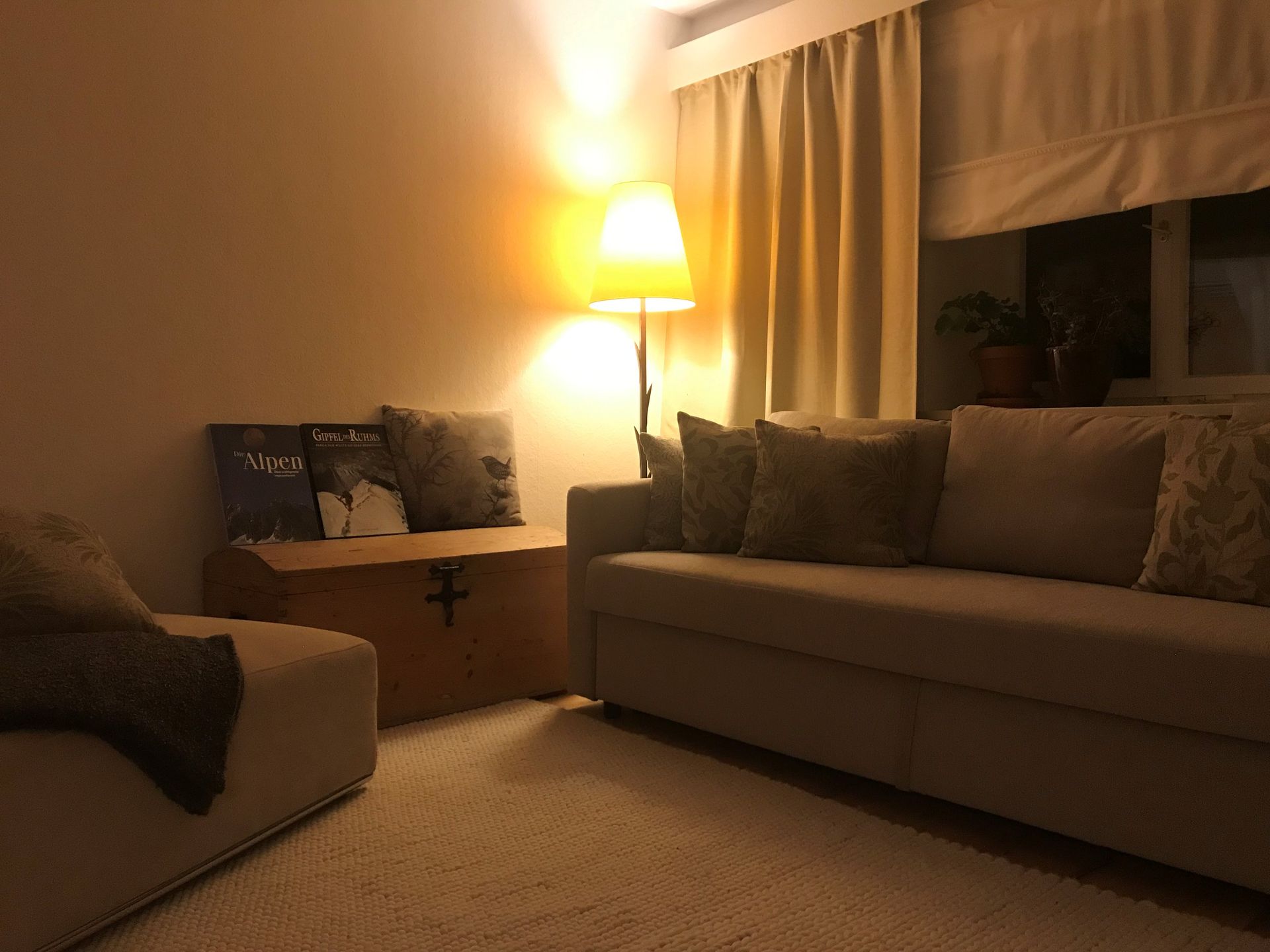 Gemütliches Wohnzimmer mit hellbraunem Sofa, Holztruhe und Stehlampe, die warmes Licht spendet.