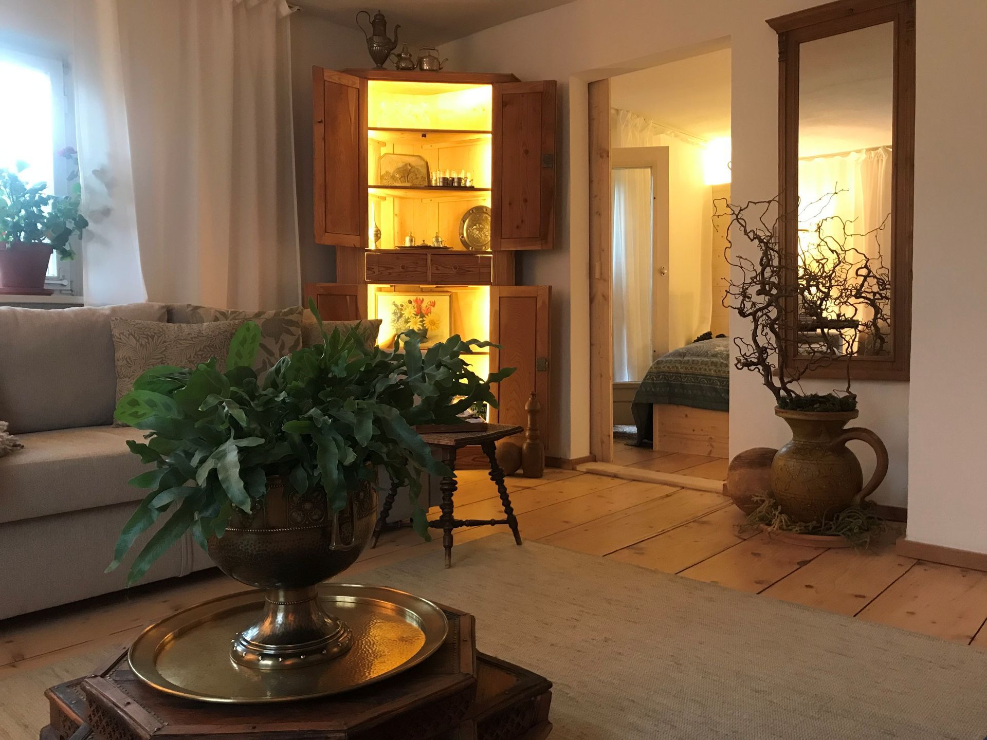 Gemütliches Wohnzimmer mit Holzmöbeln, Pflanzen und Blick in ein Schlafzimmer mit Spiegel.