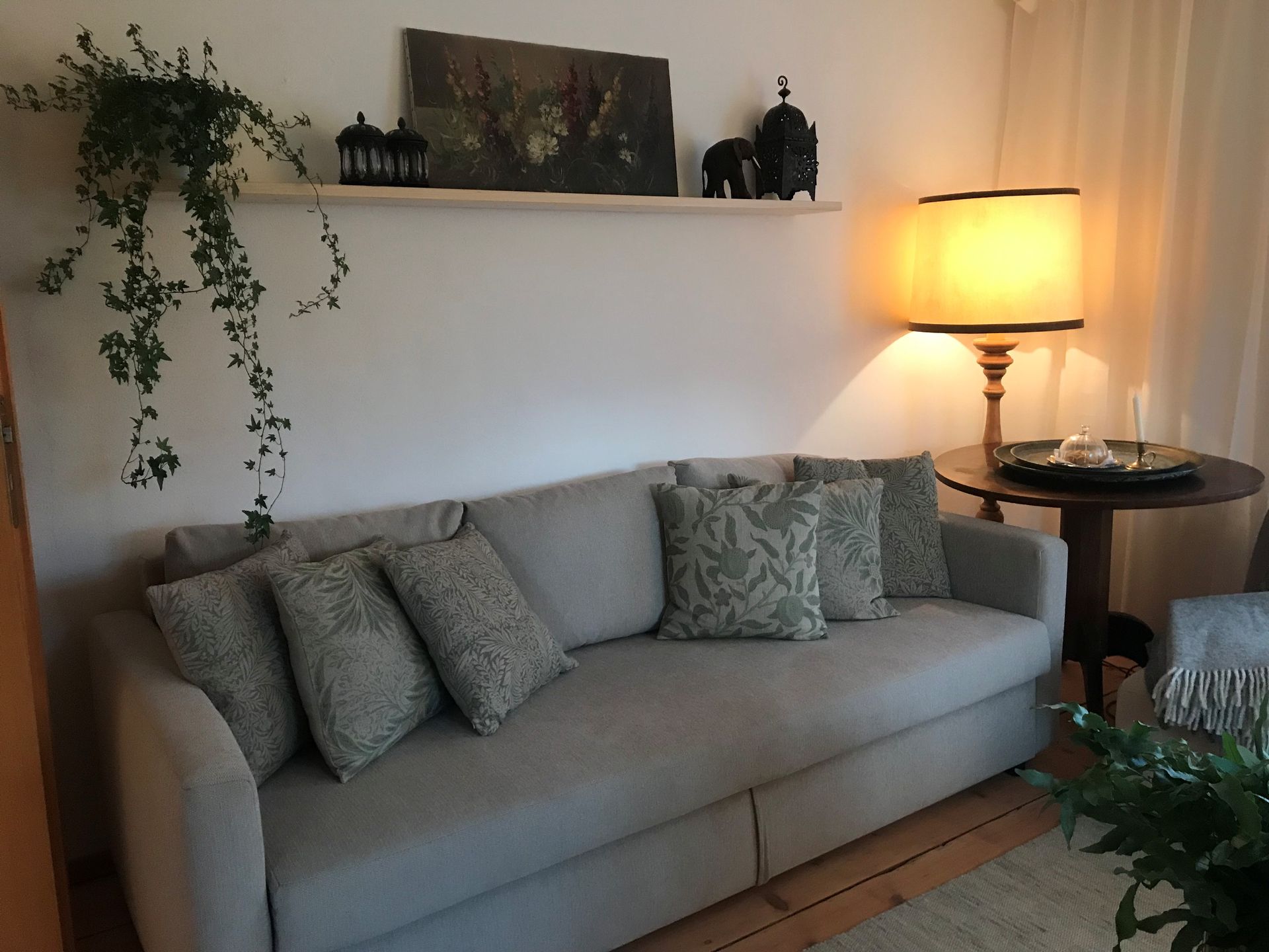 Wohnzimmer mit grauem Sofa, Kissen, Lampe und Regal mit Dekoration.