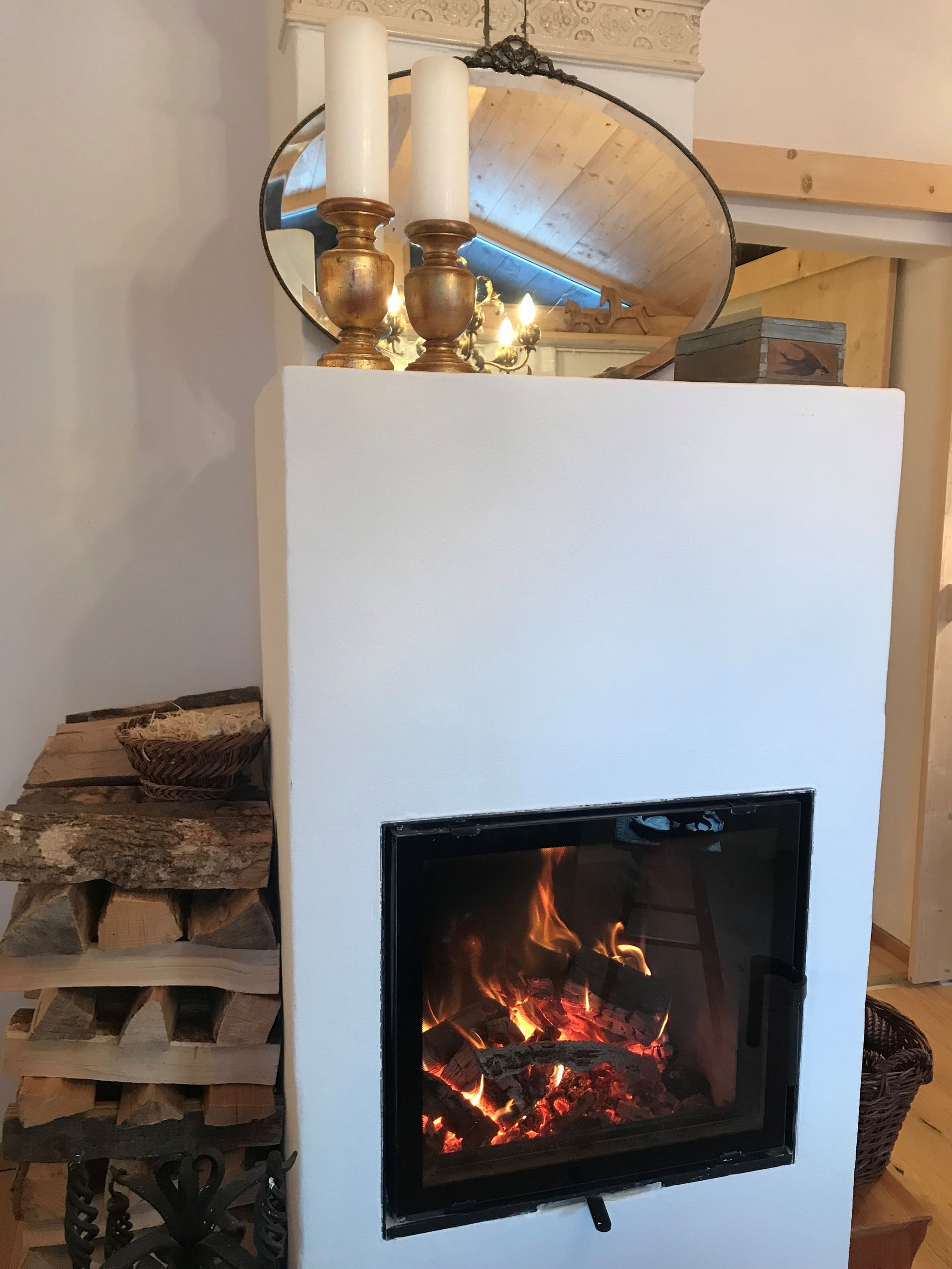 Weißer Kamin mit brennendem Feuer, Kerzen und einem Spiegel darauf, seitlich gestapeltes Brennholz.