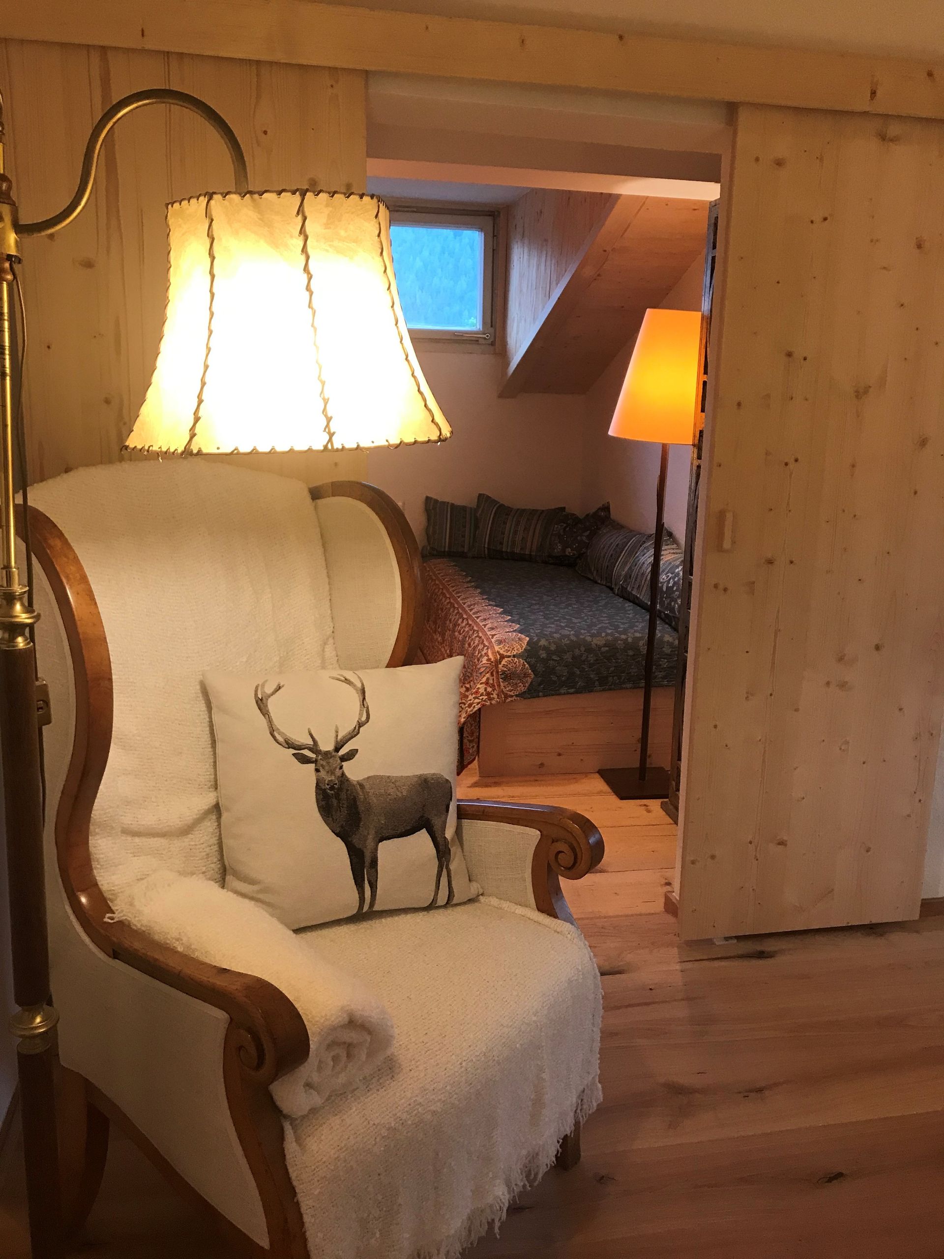 Zimmer mit Sessel, Hirschkissen, Stehlampe und Durchgang zu einem Schlafzimmer mit Bett und Lampe.