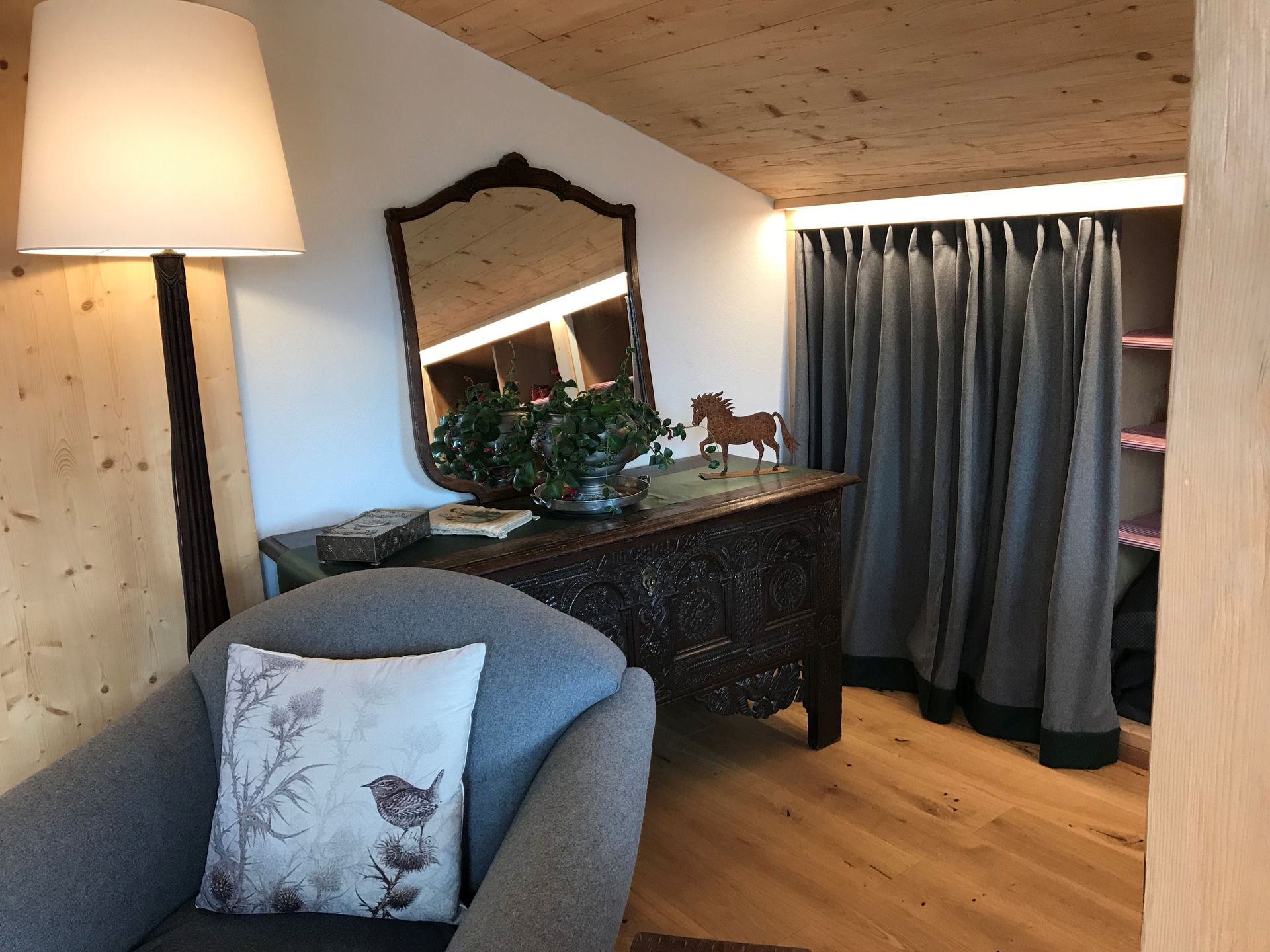 Gemütliches Zimmer mit grauem Sessel, geschnitzter Holztruhe, Spiegel, Lampe und Vorhängen.